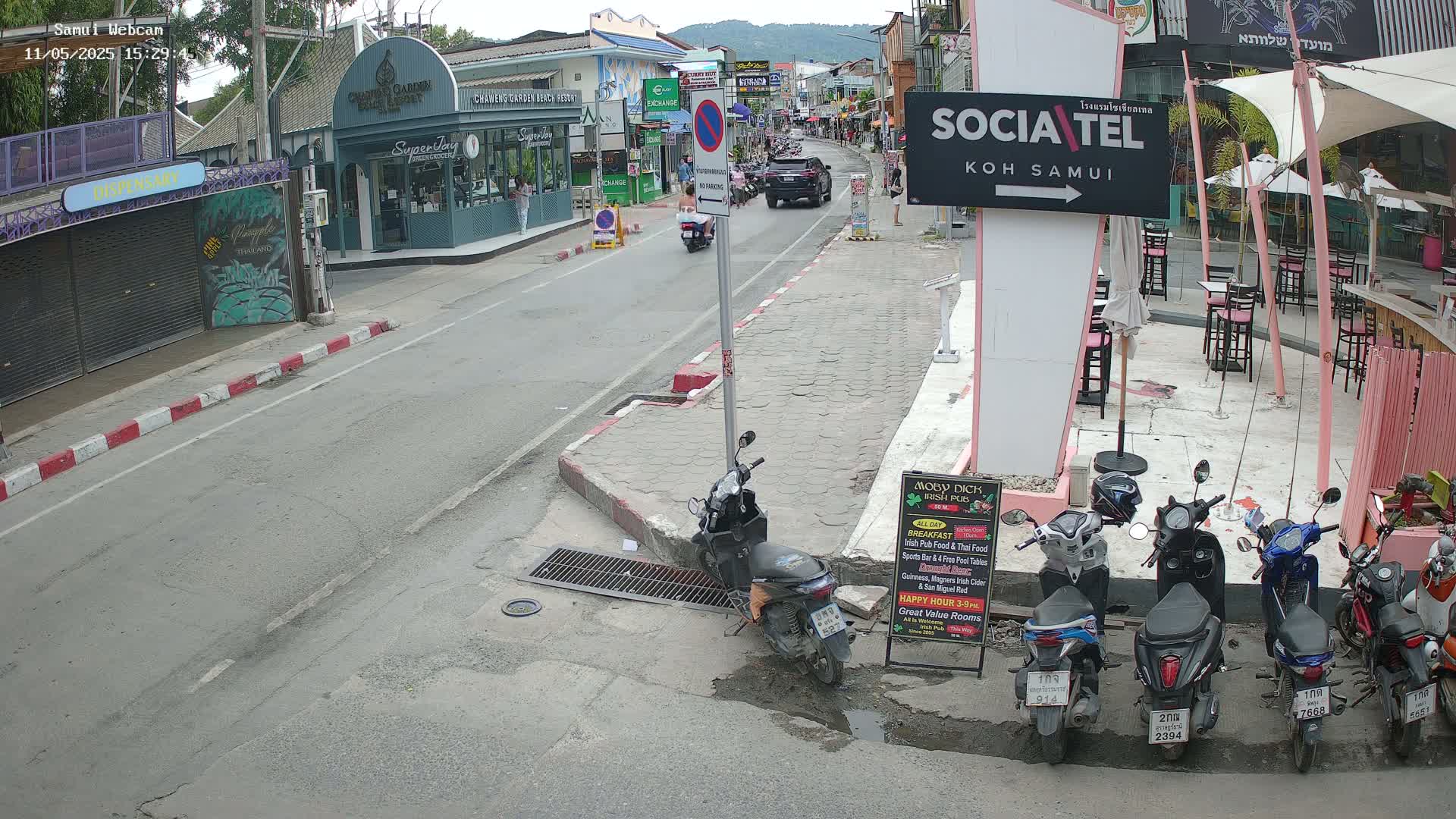 A street scene shows several parked motor scooters and a car driving away on a sunny day, with various shops and restaurants lining the street.