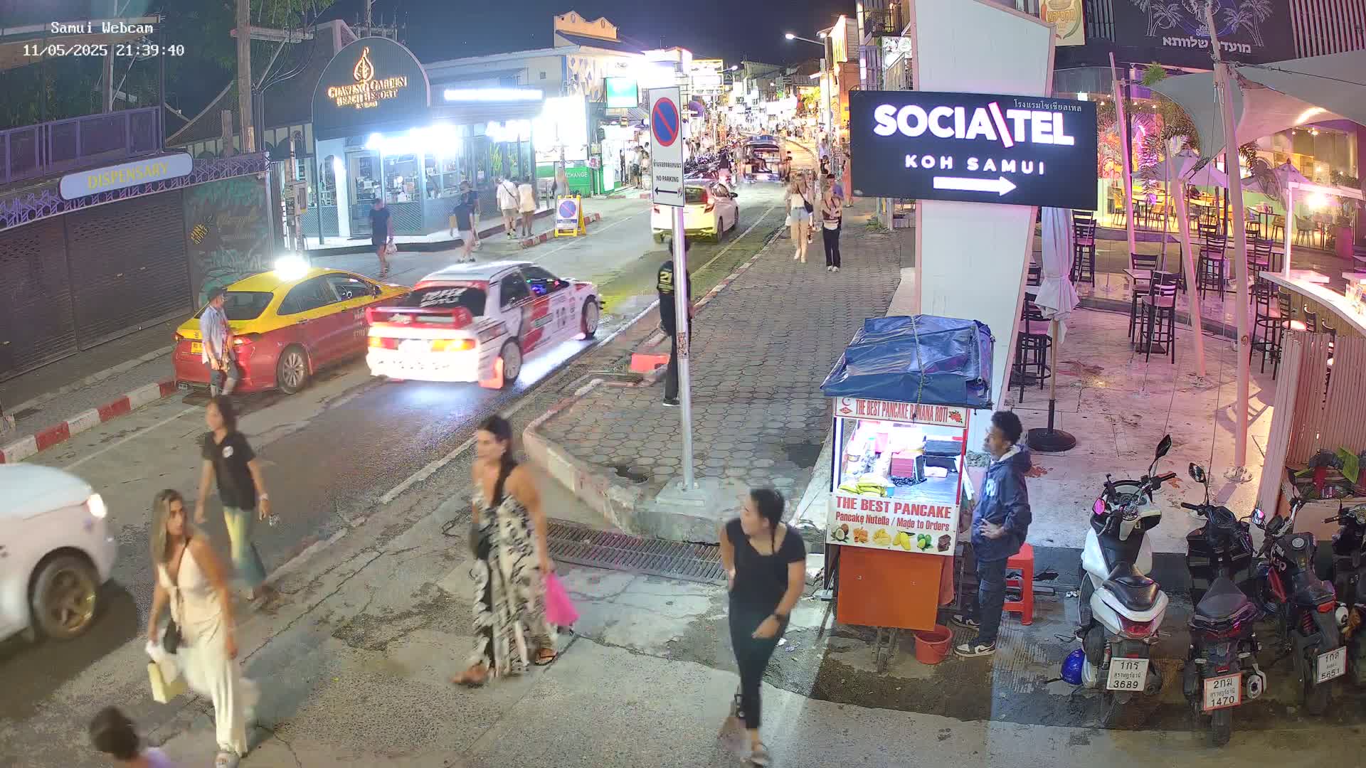 At night, under clear skies, several people stroll along a busy street lined with shops and restaurants, with cars and motorbikes parked along the sidewalks.