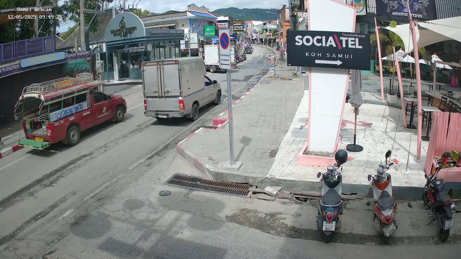 A sunny street scene shows a red songthaew, a silver pickup truck, and three parked motorbikes near a cafe.