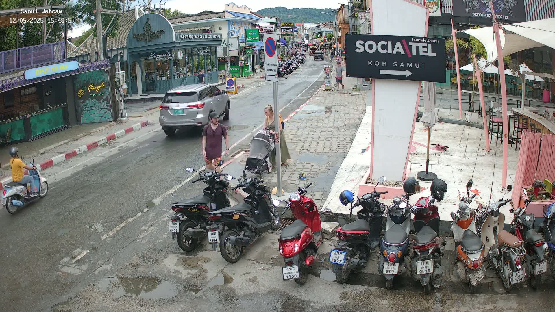 A street scene shows a couple with a baby carriage and several parked motorbikes on a slightly wet road, with a person on a motorbike driving away from the camera.