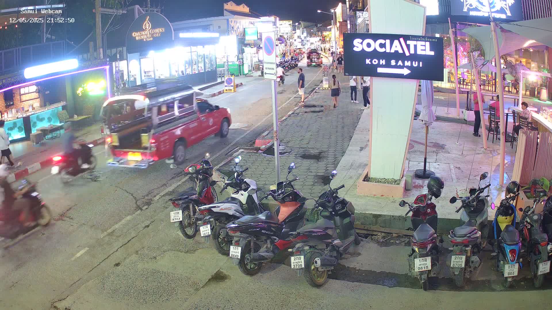 A nighttime street scene shows parked motorcycles, a red truck, and pedestrians near brightly lit storefronts.