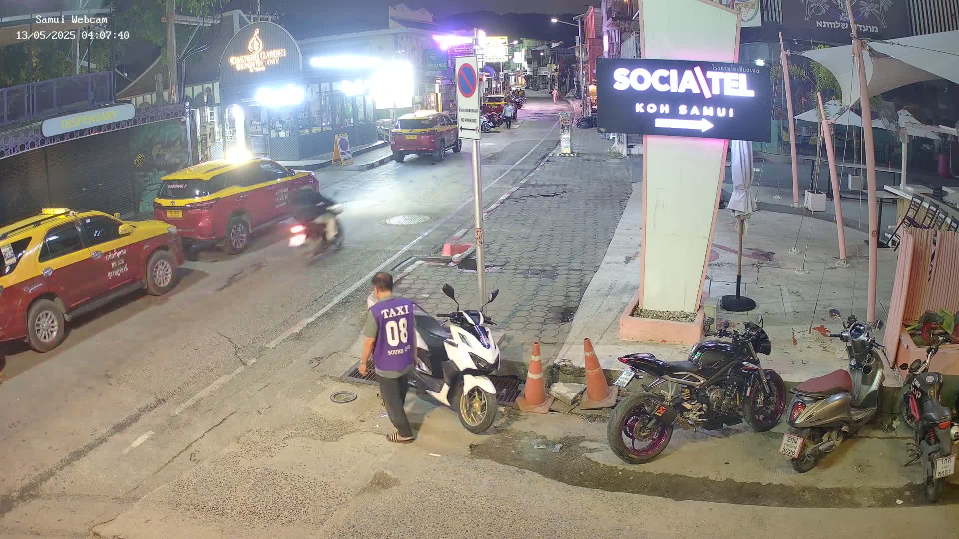 At night, a street scene shows several parked cars and motorcycles, with a person standing near a white scooter.
