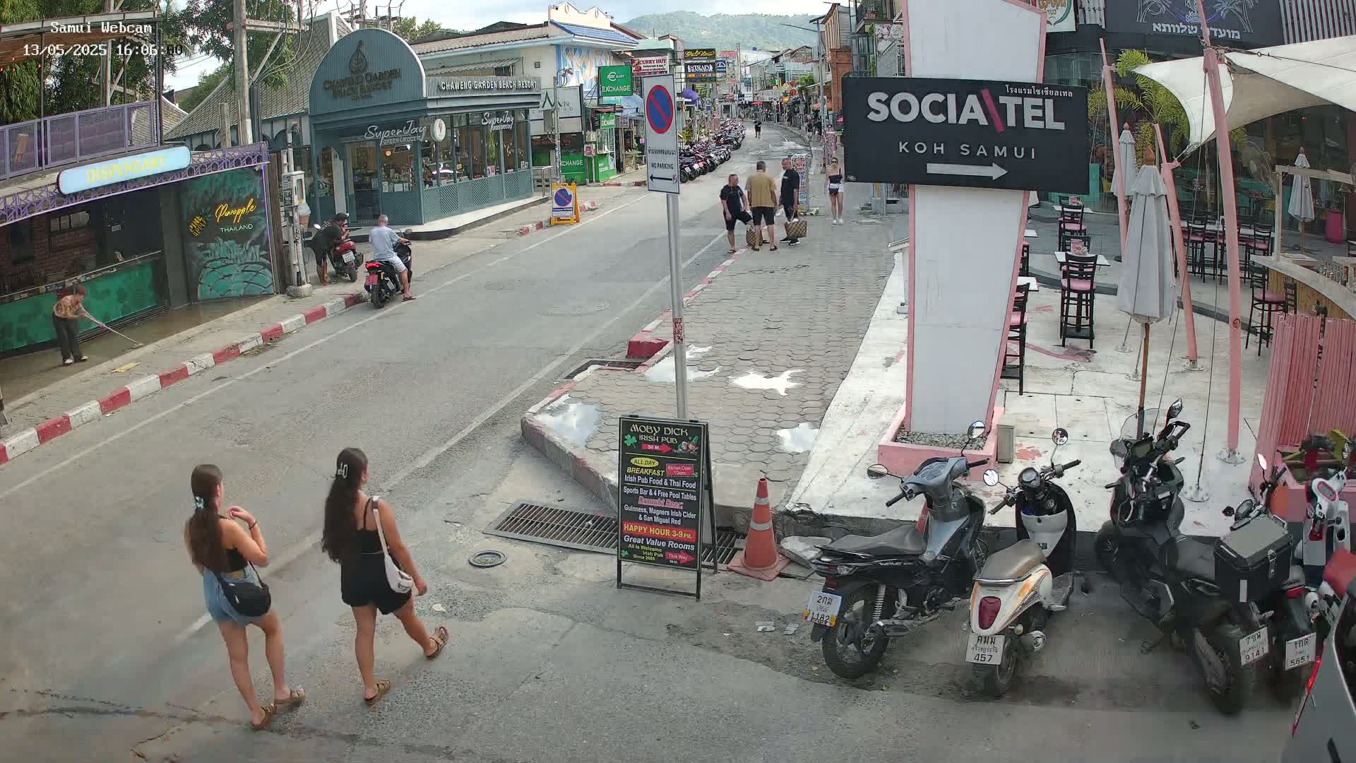 A street scene shows several people walking and motorbikes parked near outdoor seating at a restaurant under a partly sunny sky.