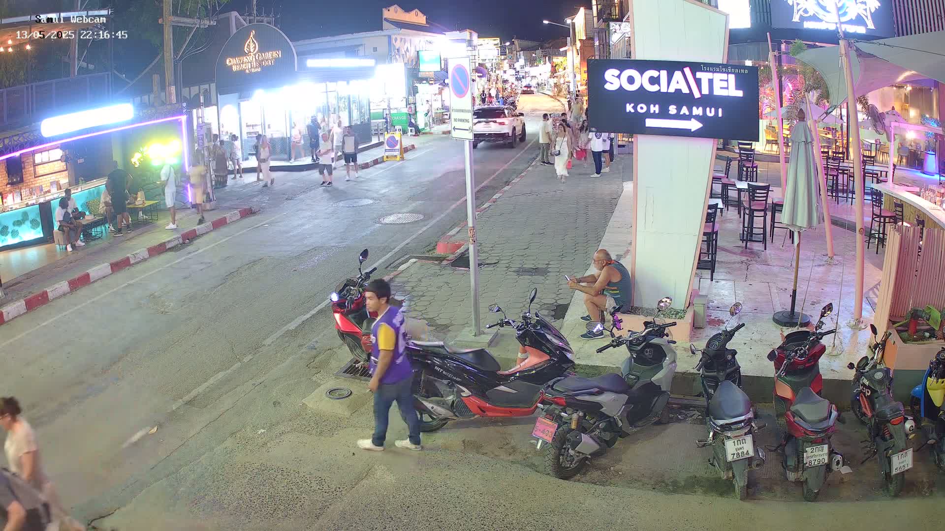 At night, a street scene shows several people walking and standing near parked motorbikes and outdoor seating areas under ambient lighting.