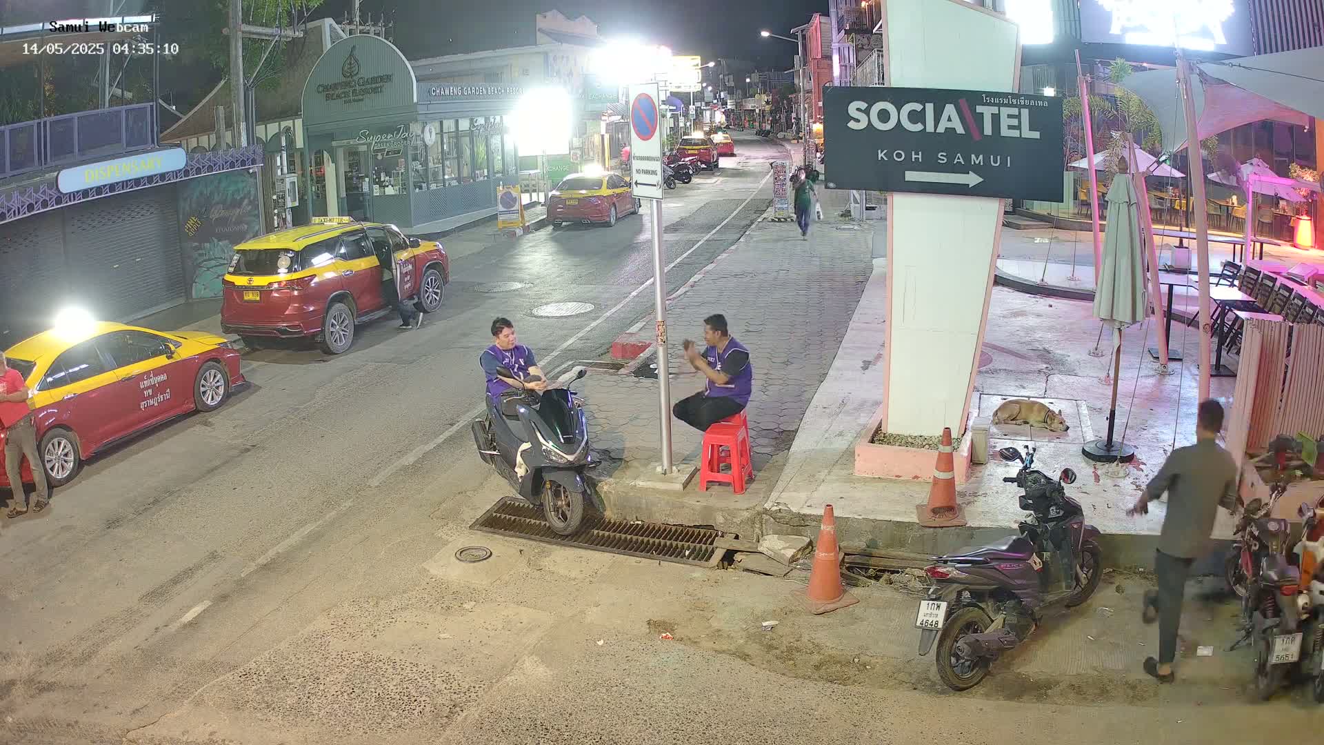 At night, on a street lined with buildings, several people stand around parked taxis and motorbikes, with one person sitting on a stool near a scooter.
