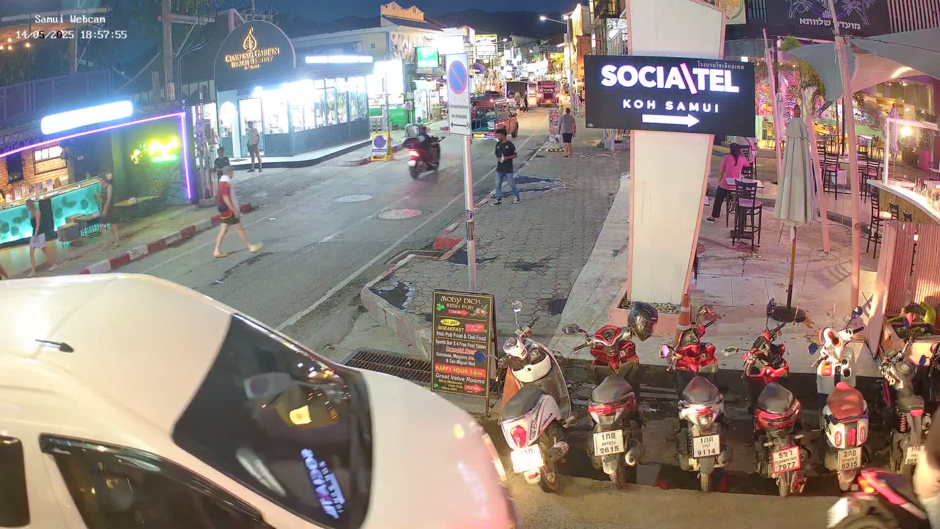 A nighttime street scene shows pedestrians and vehicles, including several parked motorbikes, under the lights of various businesses.