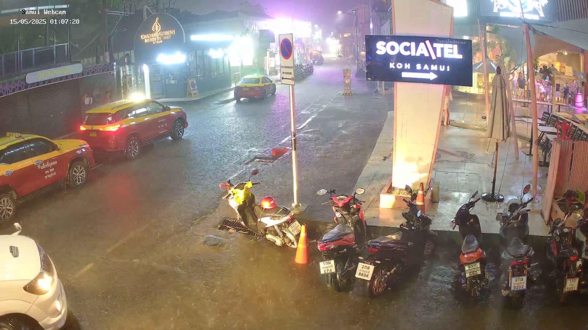 A rain-soaked street at night shows several cars and numerous parked motorcycles, with some standing water around the vehicles.