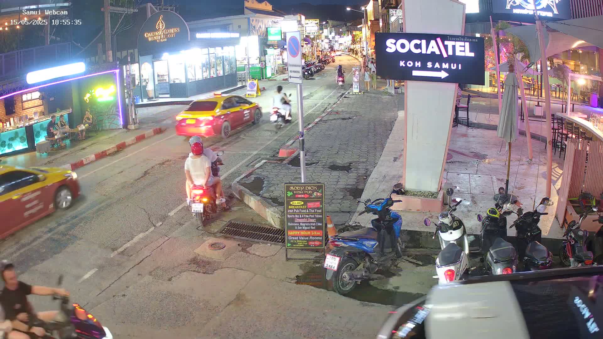 A nighttime street scene shows several people on motorbikes and in cars, parked and moving, under mostly clear skies.