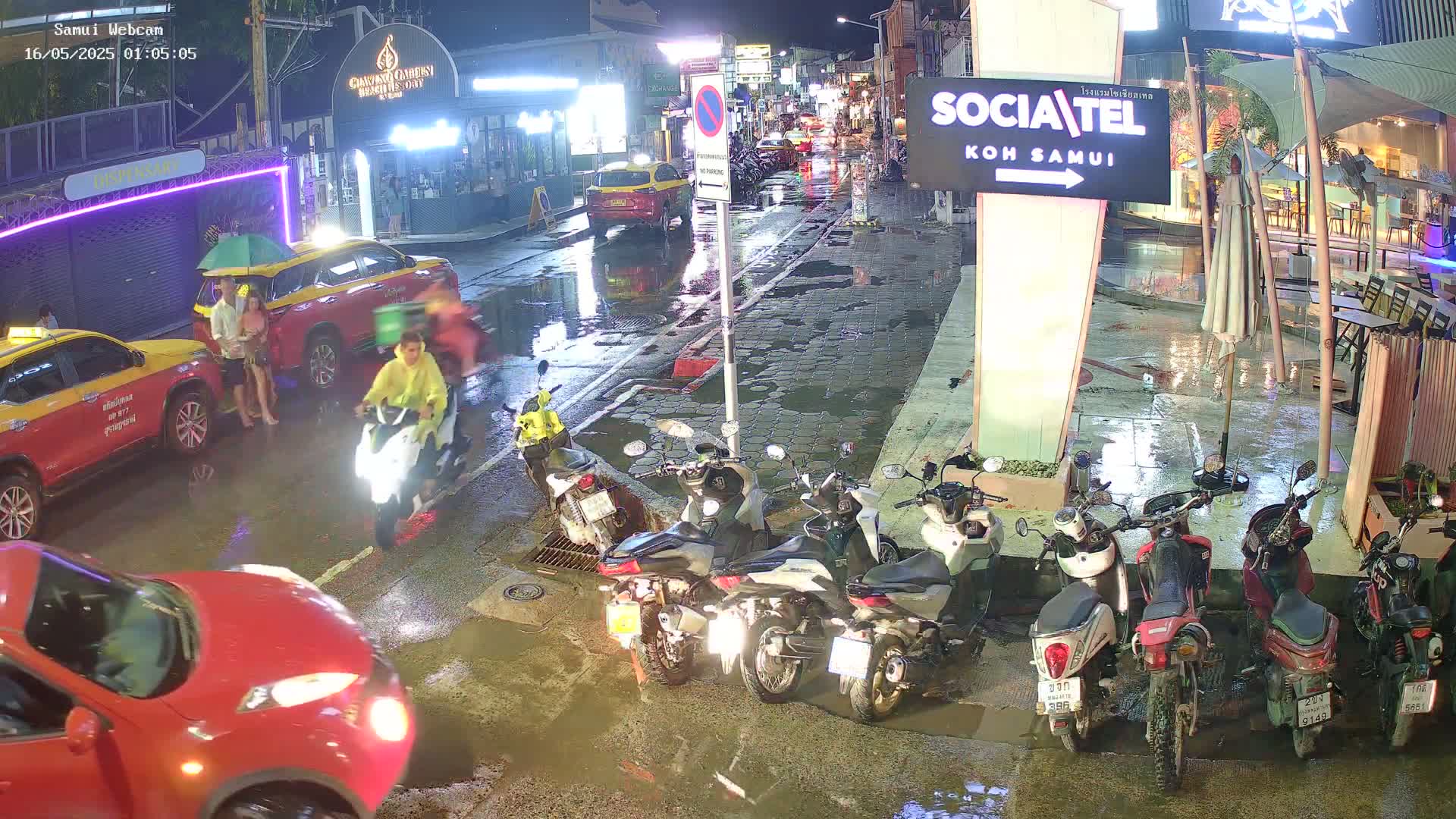 A rain-slicked street at night shows several parked motorcycles, two taxis, and pedestrians, with a person riding a scooter in the middle of the road.