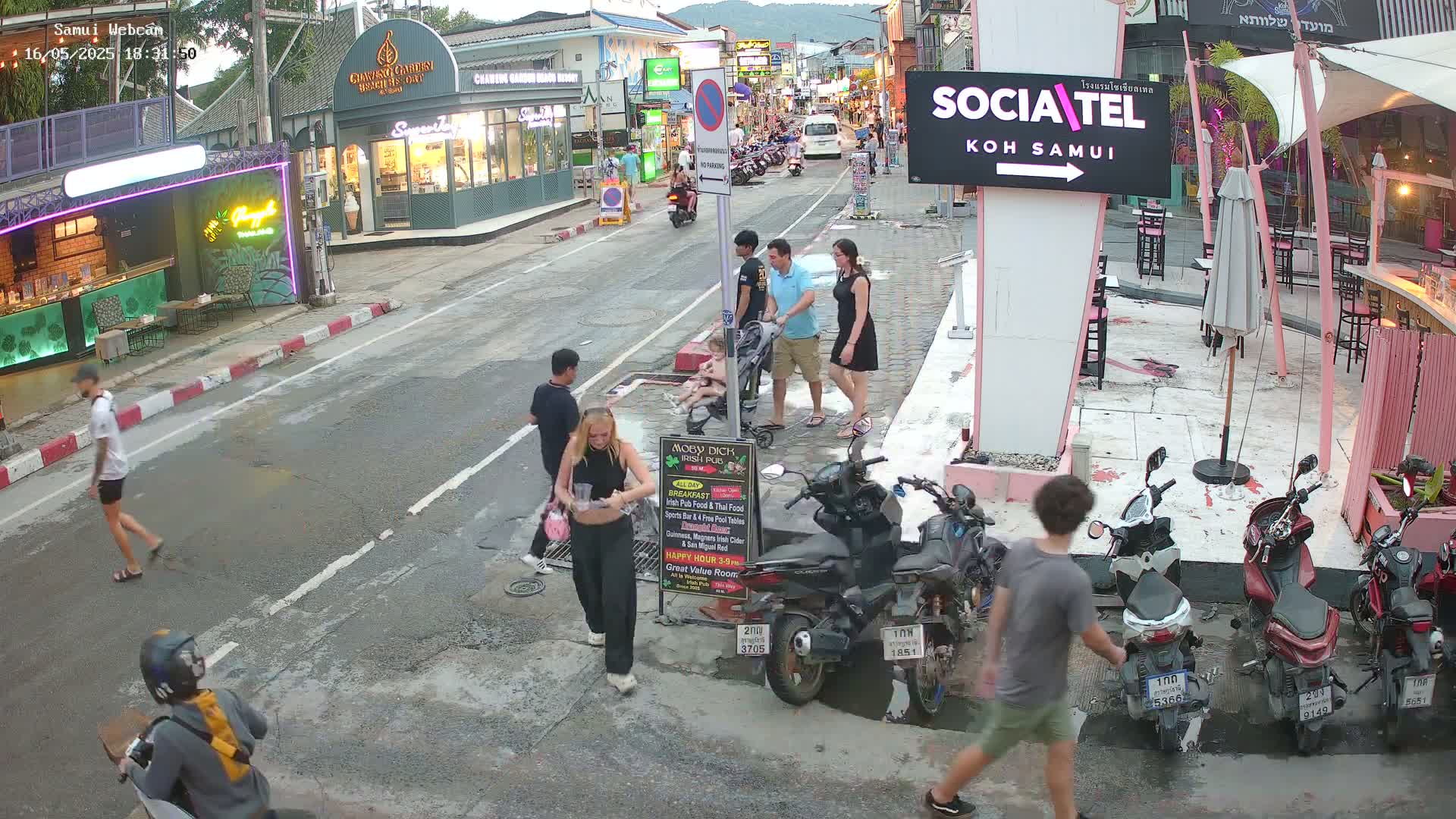 A street scene at dusk shows several people walking, some with a stroller and others on motor scooters, parked along the side of the road near shops and restaurants.