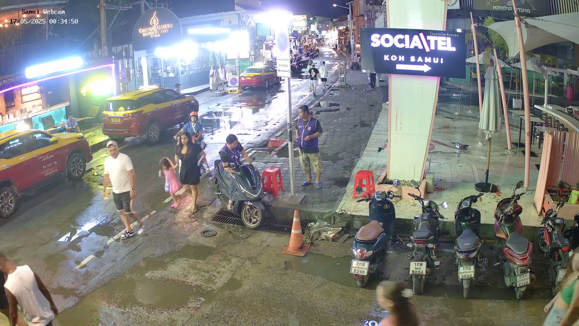 A nighttime street scene shows pedestrians and several parked motorbikes on a wet, paved road, with cars parked along the side of the street and buildings in the background.