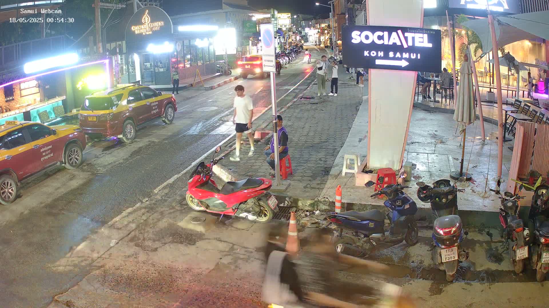 At night, several people walk and stand near parked cars and numerous motorbikes on a wet street next to some businesses.