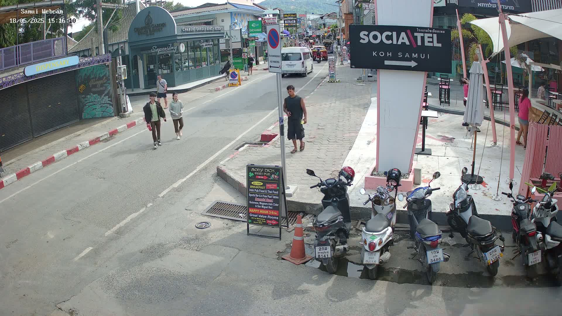 A sunny street scene shows several people walking and a few motorbikes parked, alongside buildings and outdoor seating areas.
