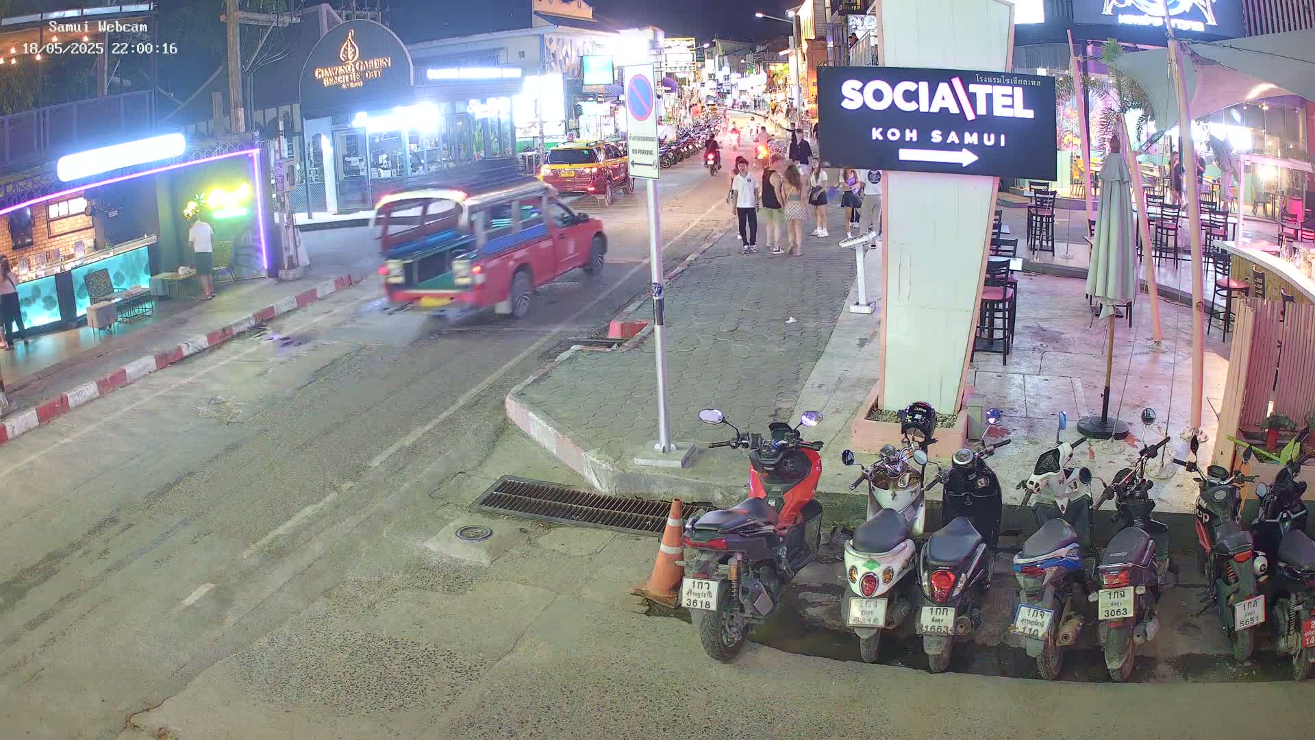 A nighttime street scene shows a red truck turning a corner, pedestrians walking, several parked motorcycles, and outdoor seating areas at nearby businesses.