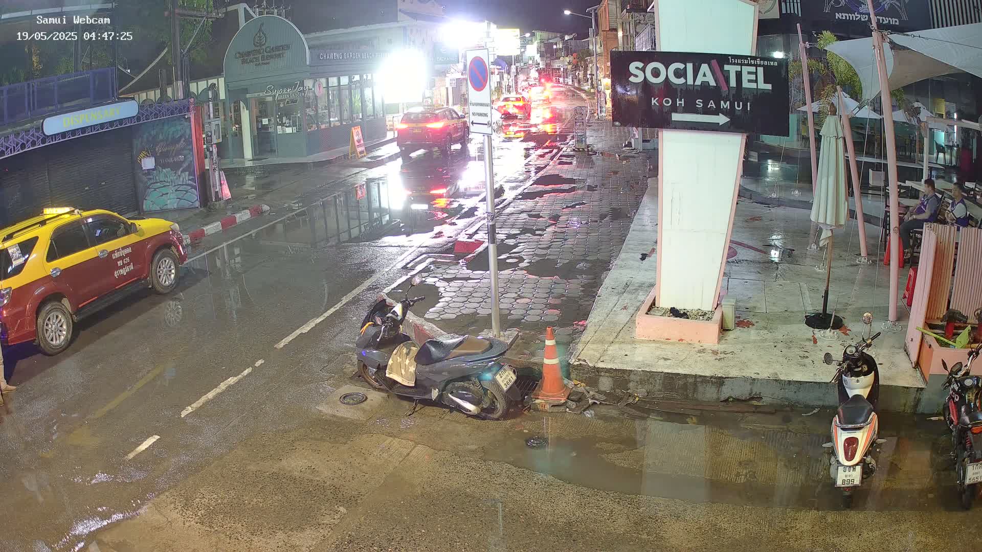 A rain-slicked street at night shows a taxi, several motorbikes, and cars parked along the side of the road, with puddles reflecting the streetlights.