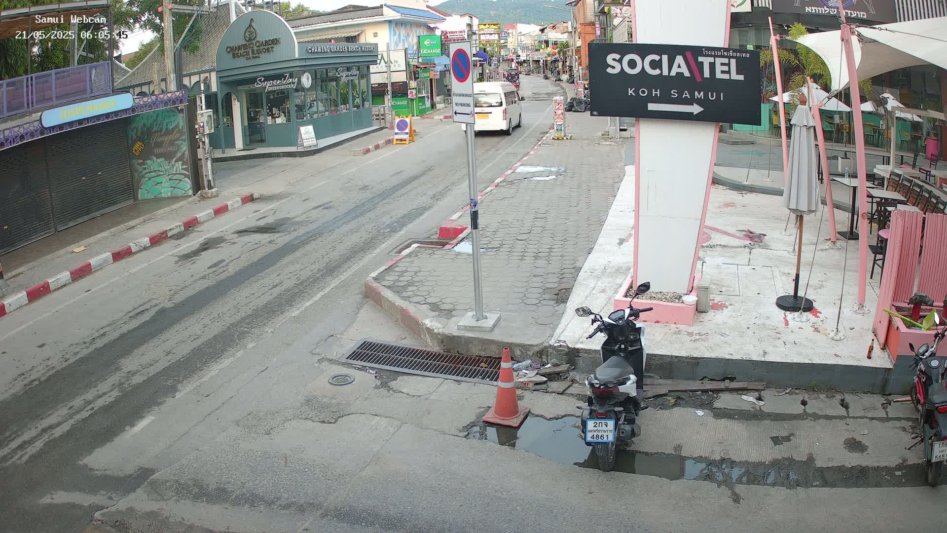 A wet street scene shows a minivan and several parked motorcycles near shops and restaurants, under an overcast sky.