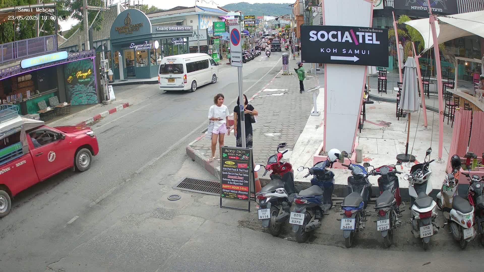A street scene shows two women standing on a sidewalk near parked motorbikes and a red pickup truck, with several shops and restaurants visible in the background under a clear sky.