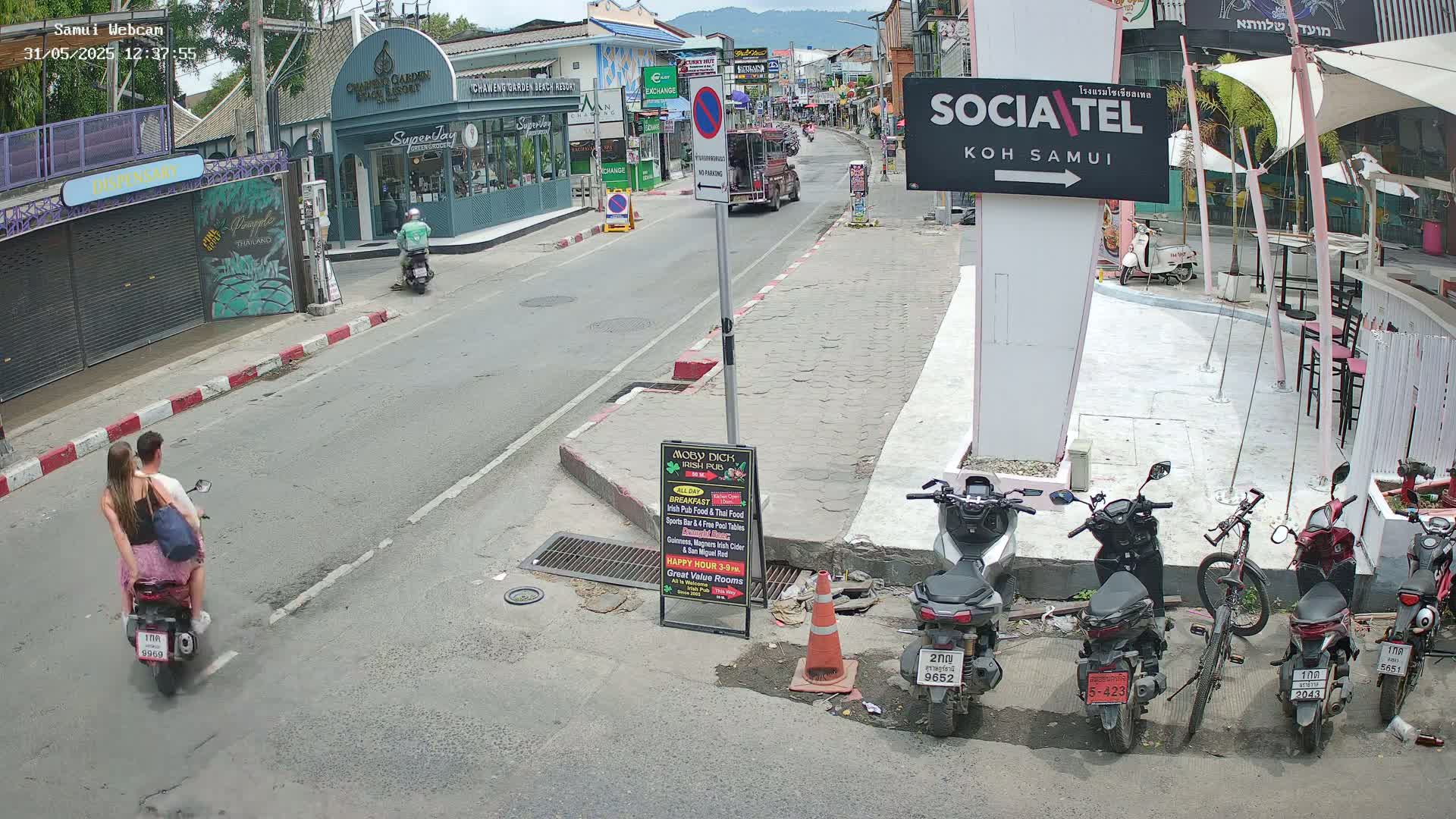 A couple rides a motorbike down a sunny street lined with shops and parked motorcycles.
