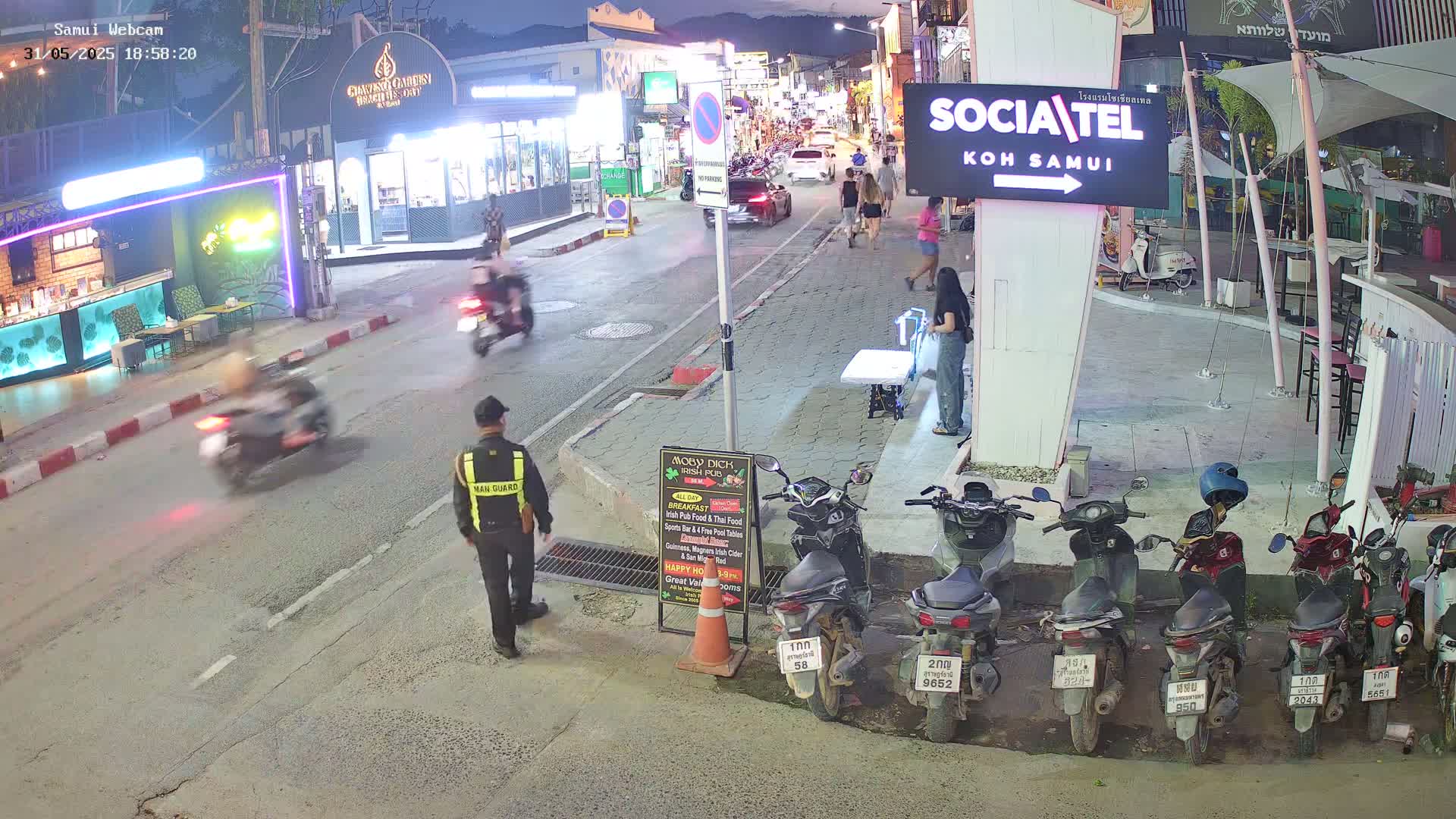A nighttime street scene shows several motorbikes moving and parked, pedestrians walking, and a security guard standing near a row of parked motorbikes.