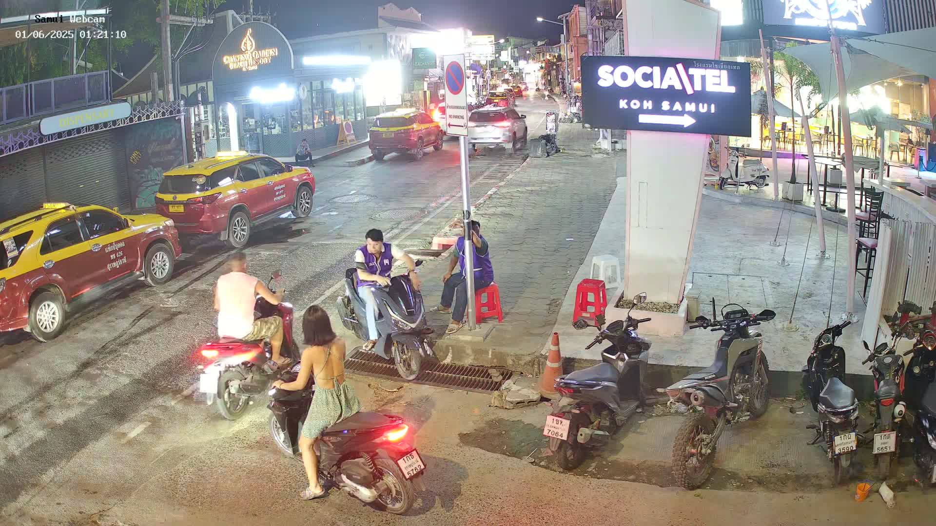 At night, several people are gathered around motorbikes and scooters parked on a street, near taxis and other vehicles; some people are sitting on scooters.