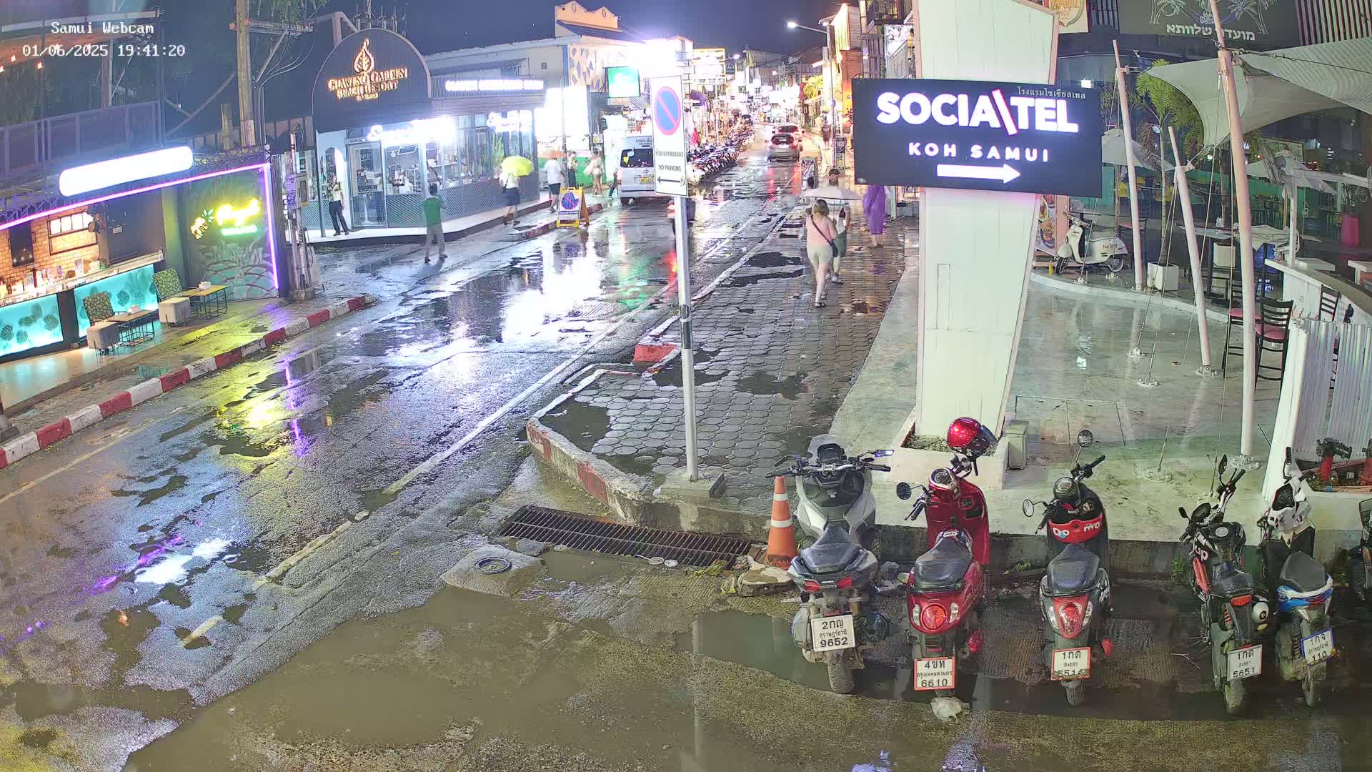 A rain-slicked street at night, showing several parked motorbikes and a few pedestrians walking past shops and restaurants.