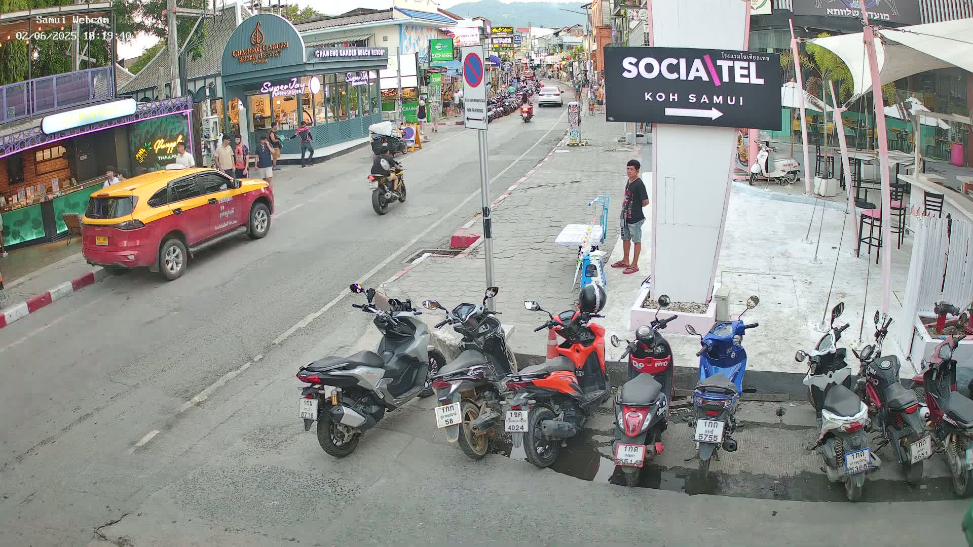 A street scene shows several parked motorbikes and a yellow and red taxi, with a man standing near a sign, under a partly cloudy sky.