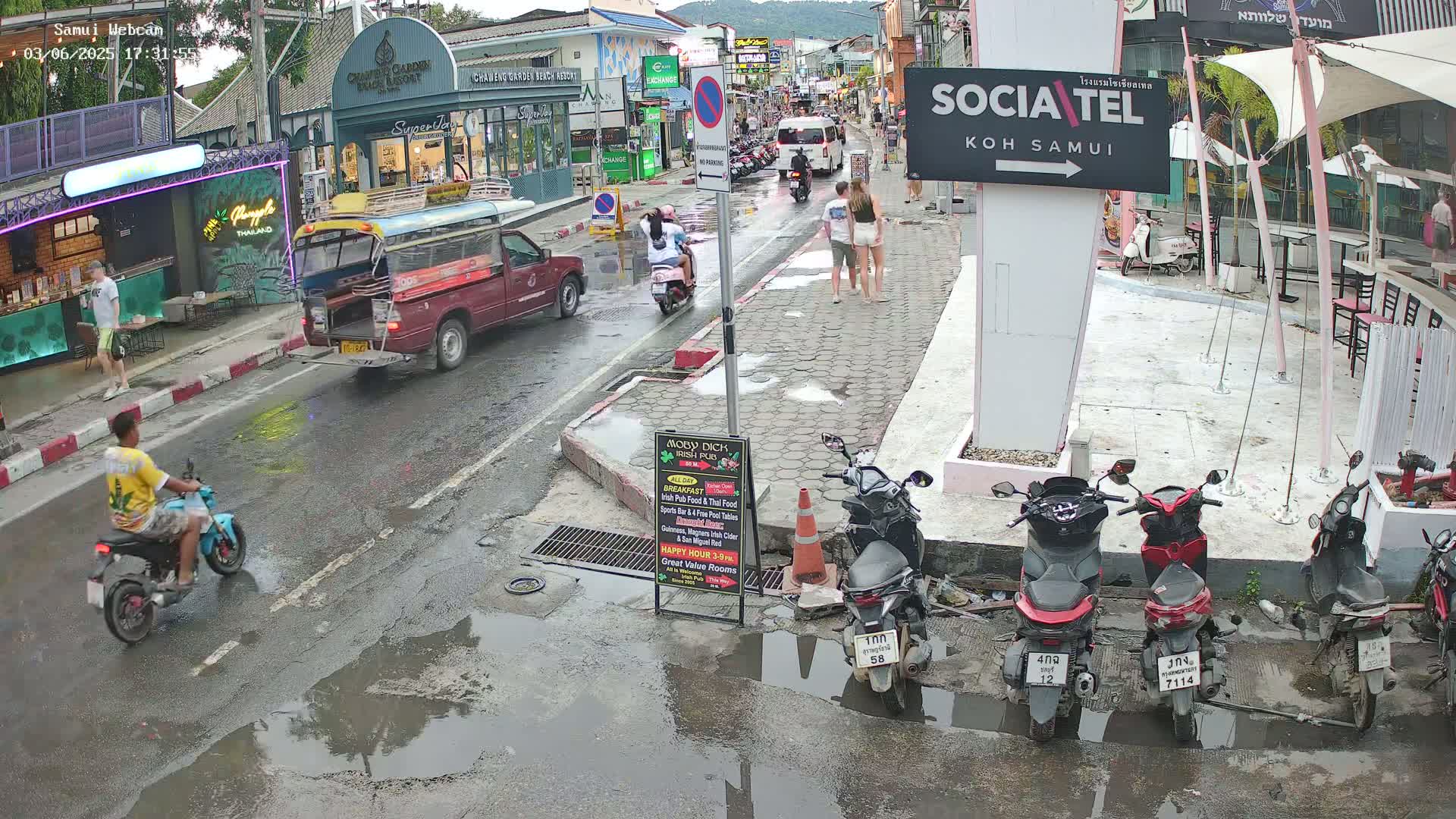 A wet street scene shows people walking and riding motorbikes and scooters past shops and restaurants, with several parked scooters and a red pickup truck.