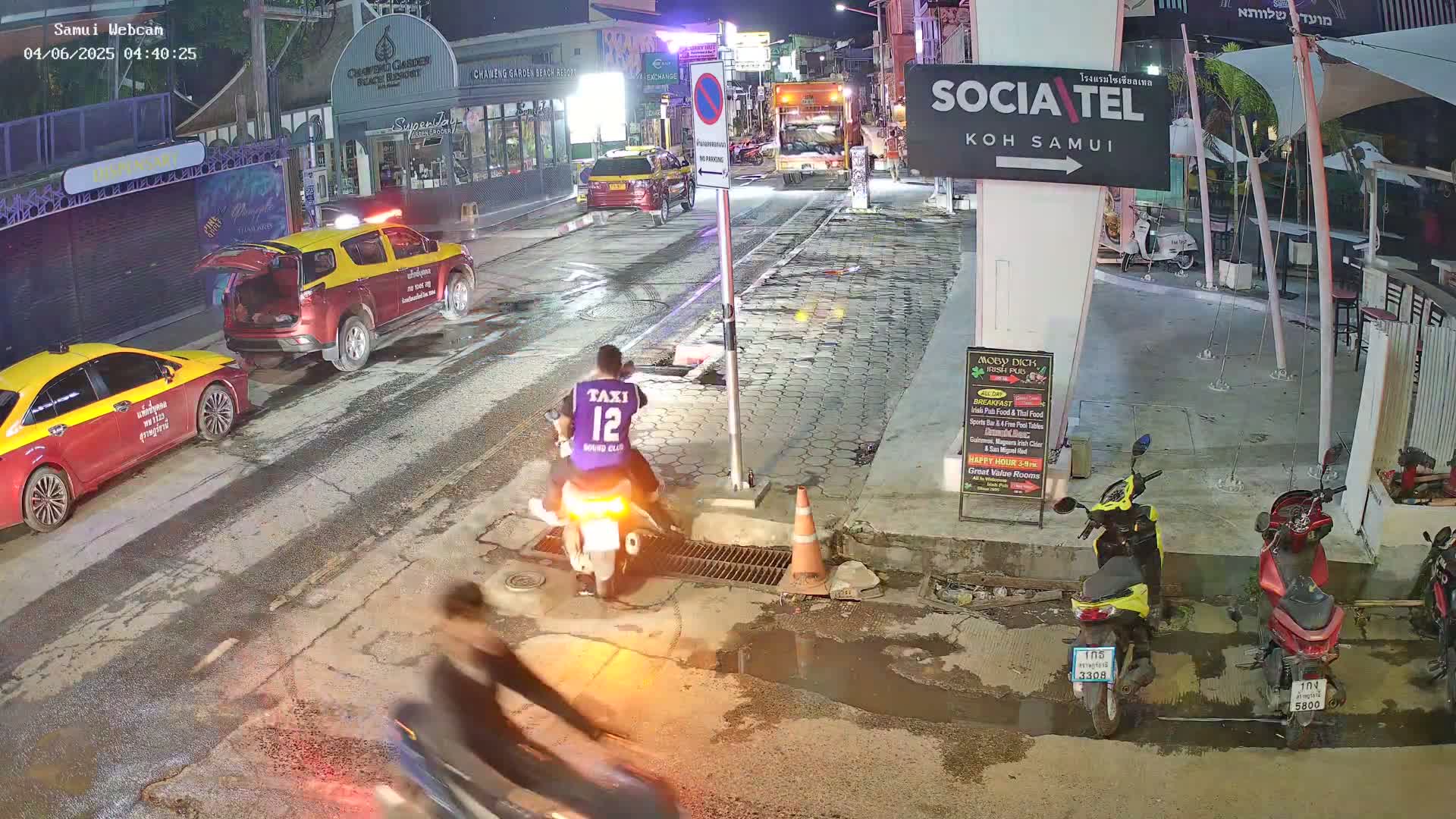 At night, on a wet street, a person sits on a motorbike, two taxis are parked nearby, and several motorbikes are parked along a building.