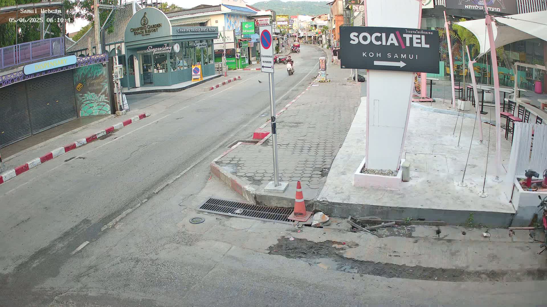 A mostly empty street scene in daytime shows a road with buildings on either side, a person on a scooter in the distance, and some minor street construction.