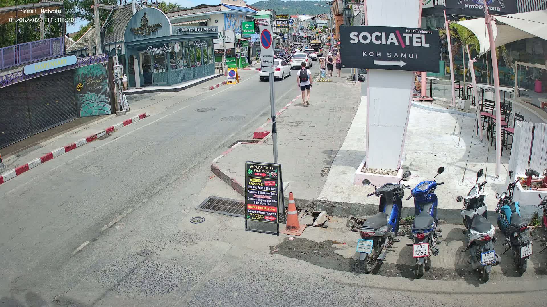 A sunny street scene shows several parked motorbikes, a person walking, and cars driving past shops and restaurants.