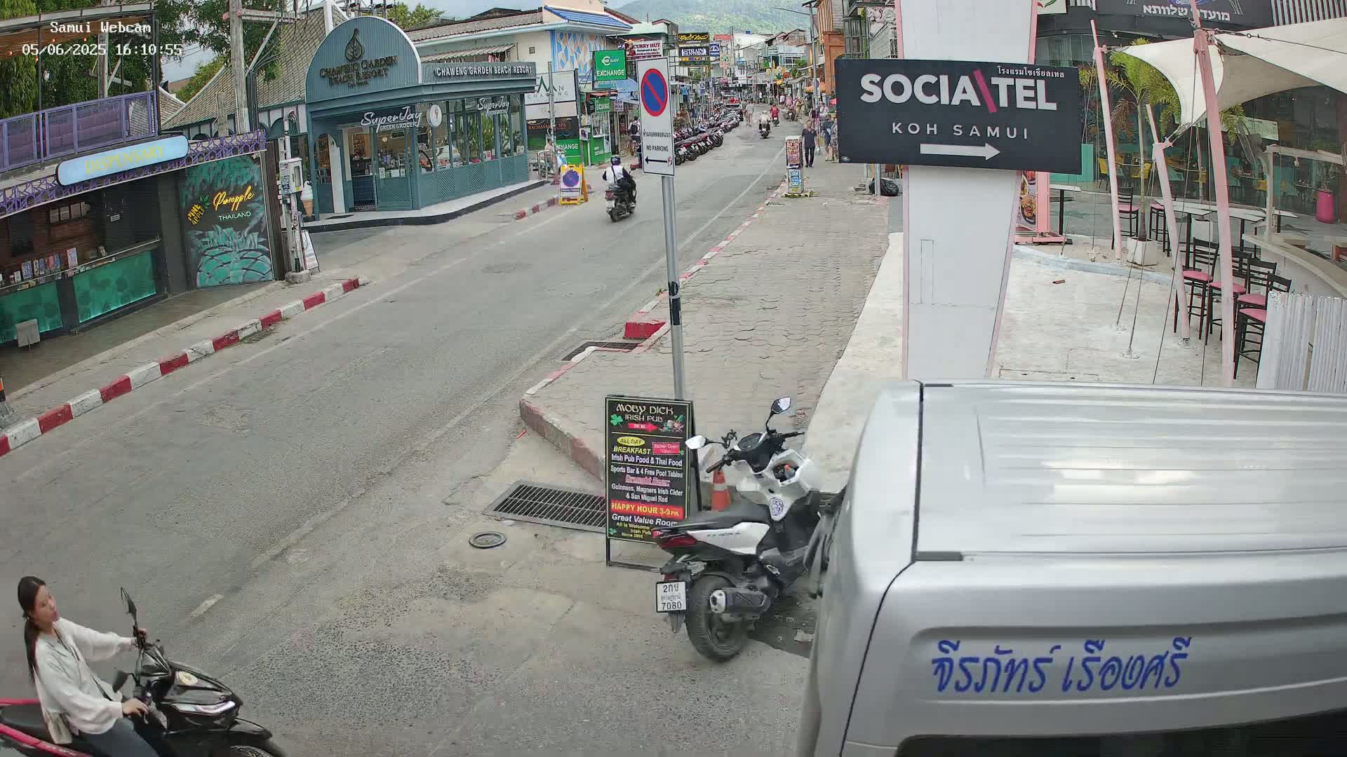 A street scene shows several parked and moving motorbikes, pedestrians, and buildings on a sunny day.