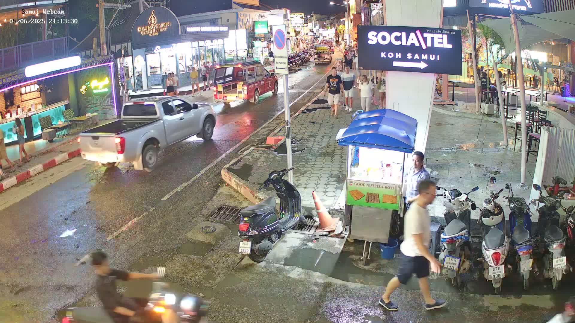 A nighttime street scene shows a wet road with several pedestrians, a pickup truck, scooters, and parked motorbikes near food stalls and shops.