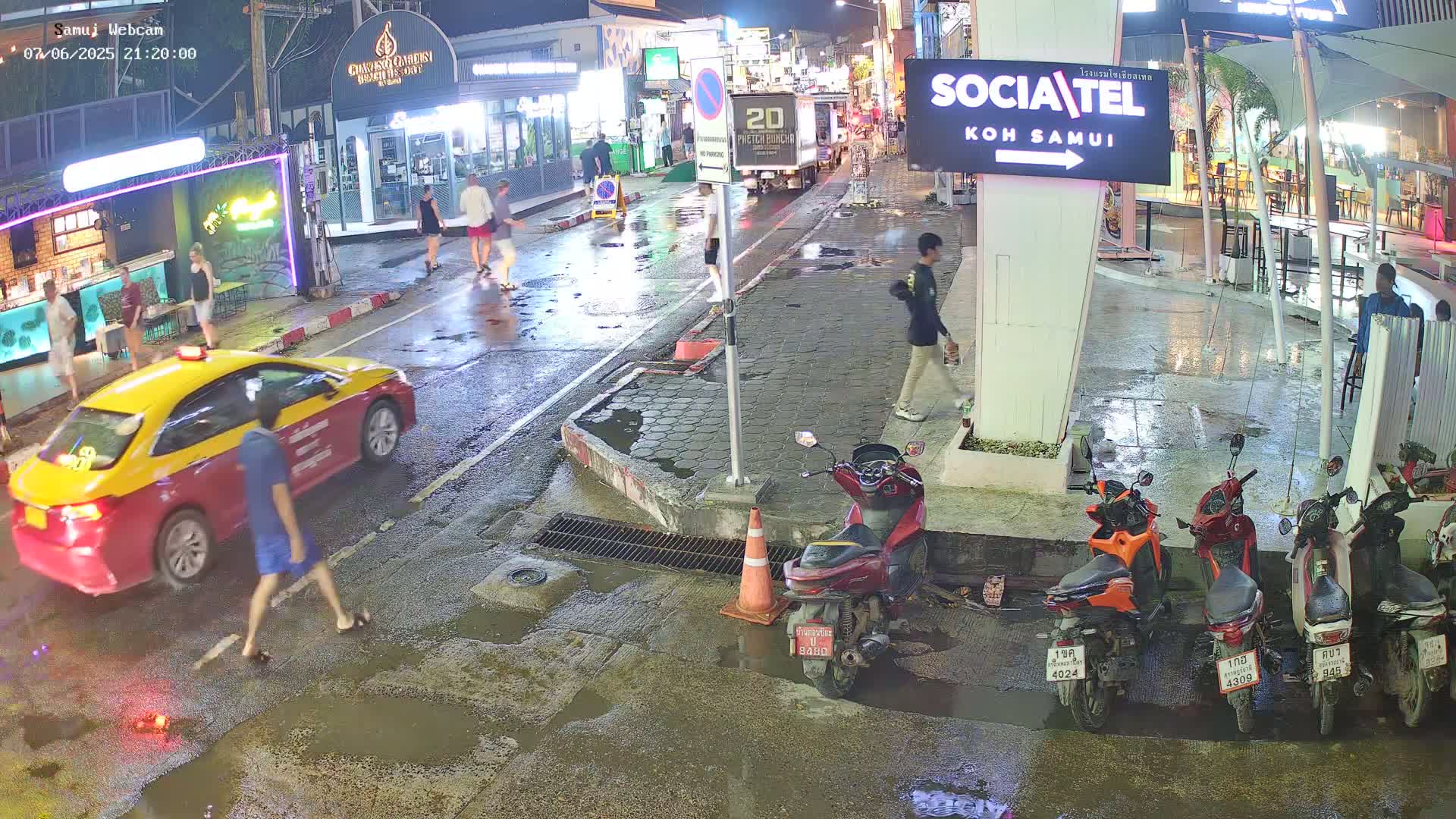 A wet, nighttime street scene shows a yellow and red taxi, pedestrians, several parked motor scooters, and some people walking near shops and restaurants.