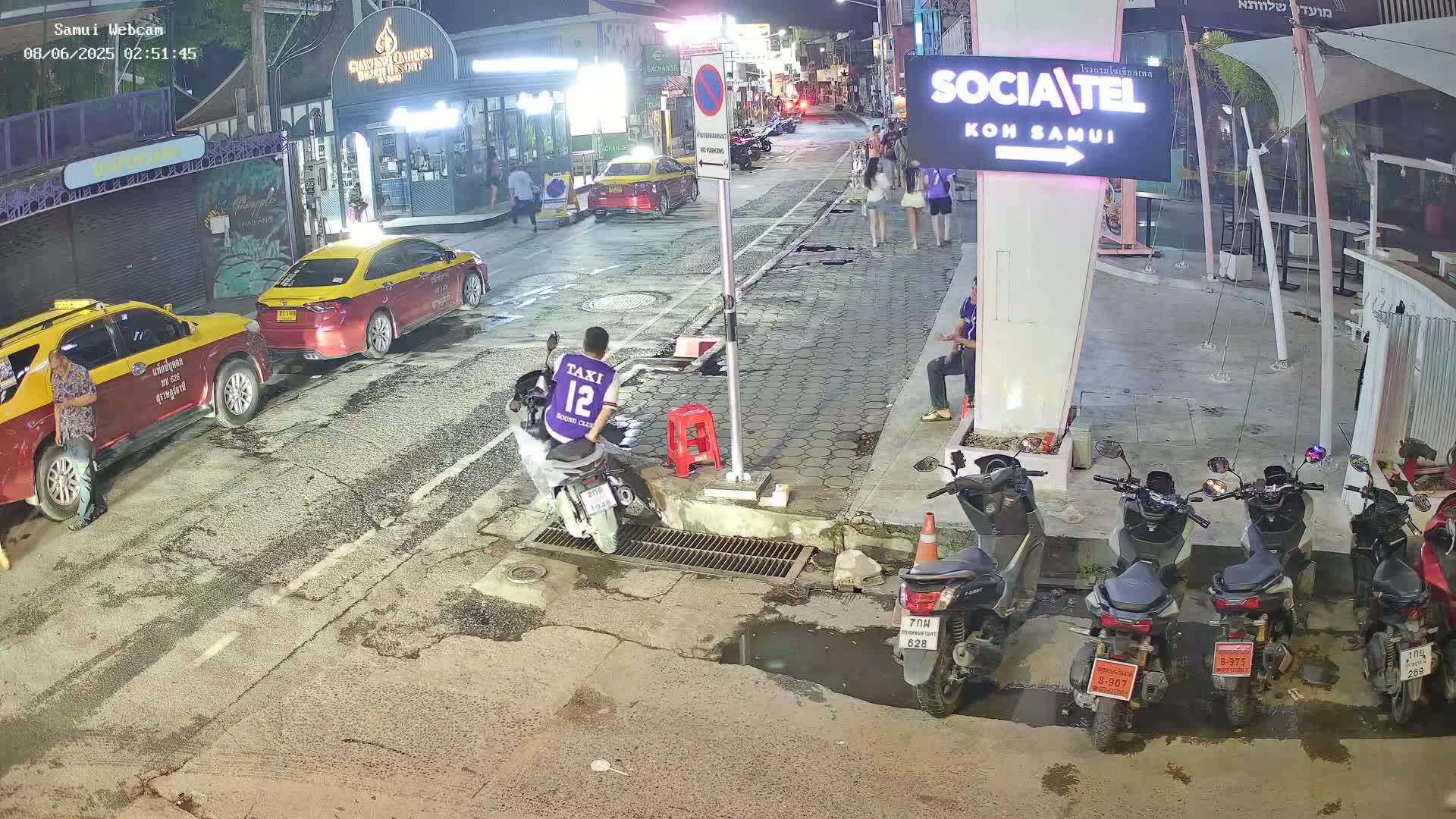 At night, a street scene shows several parked taxis and motorbikes near a building, with a person on a motorbike and a few pedestrians visible.