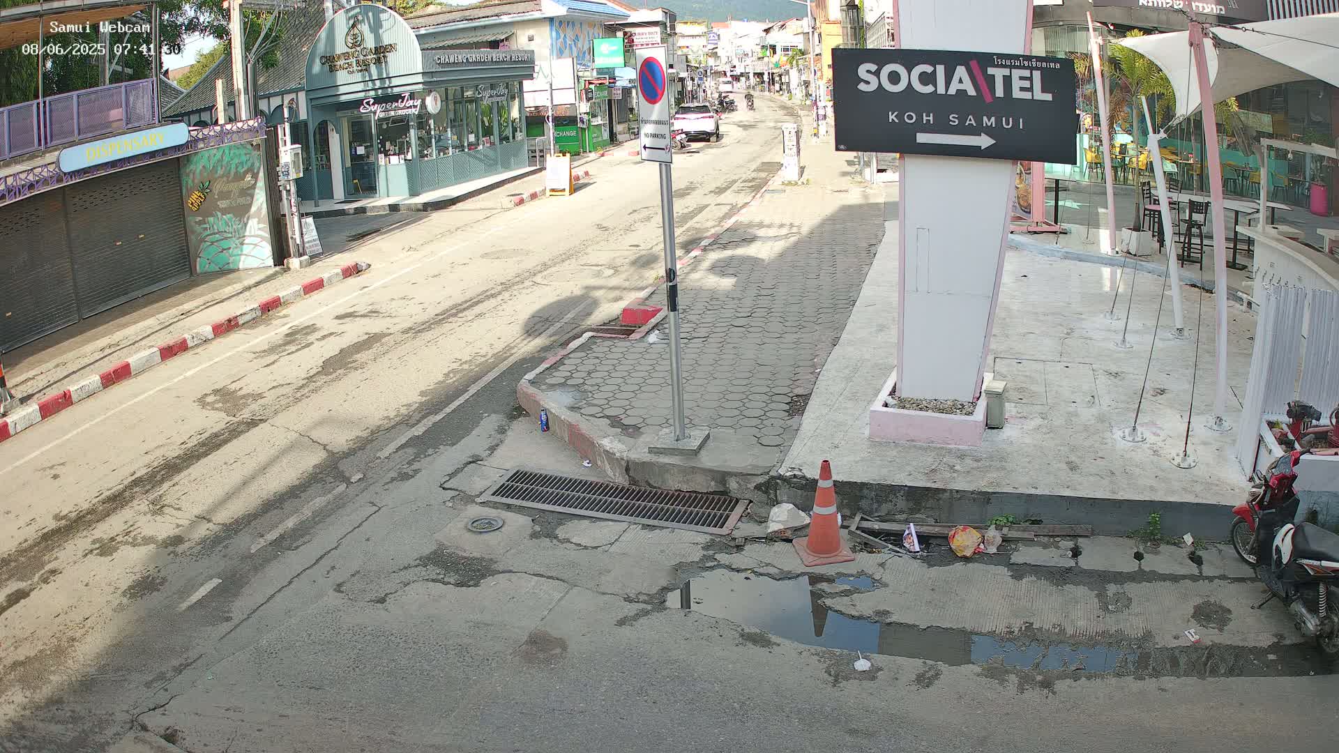 A mostly empty street scene on a sunny day shows buildings and shops lining both sides of the road, with a small puddle and a traffic cone near the curb.