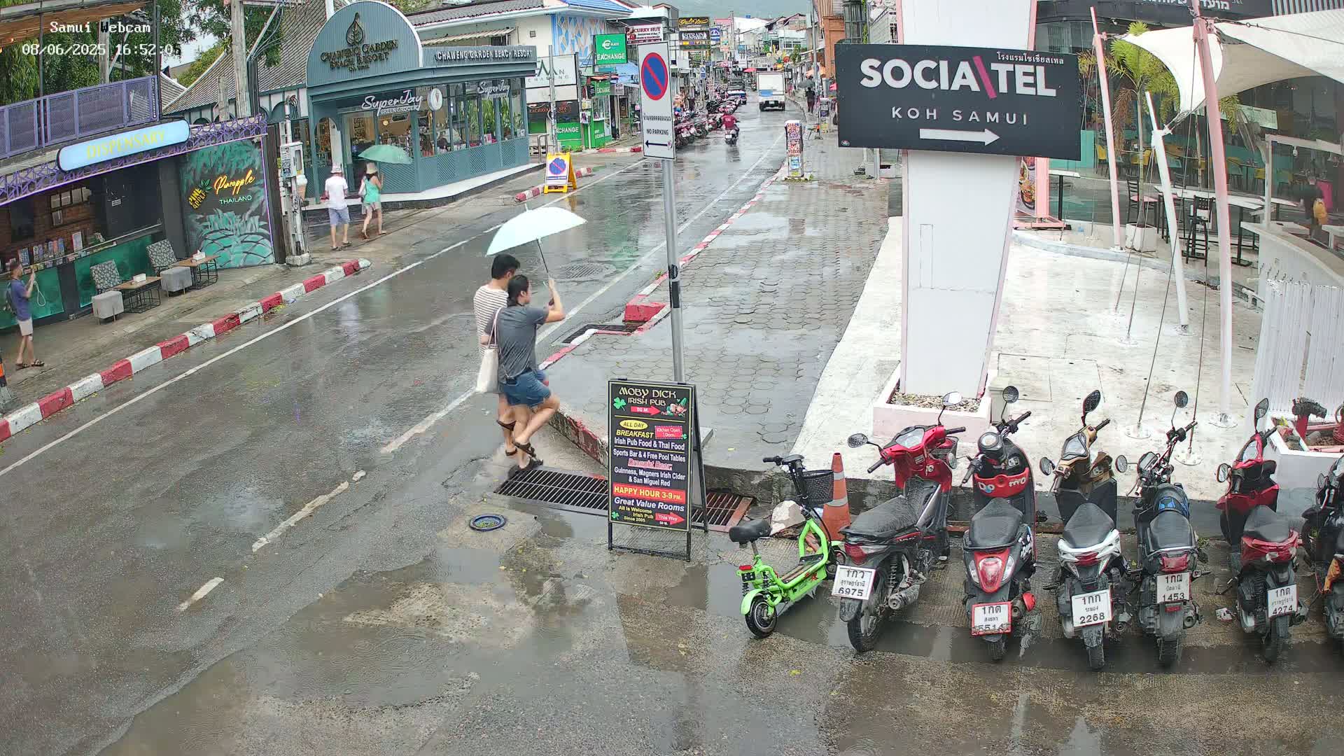 A wet street scene shows two people walking under an umbrella, several parked motorcycles, and some shops and buildings in the background under a rainy sky.