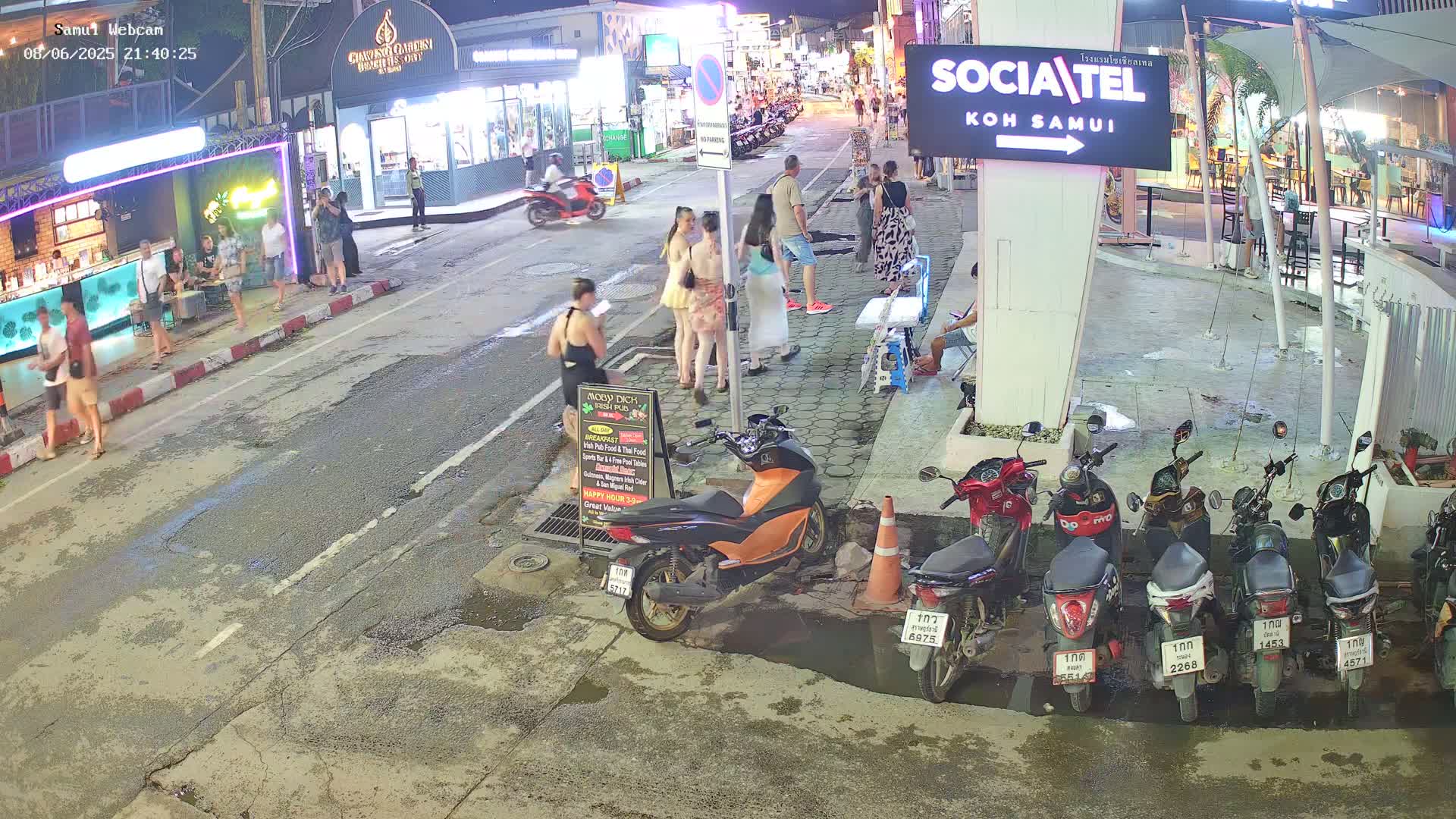A nighttime street scene shows several people walking and standing near shops and restaurants, with parked motorbikes lining the side of the road.