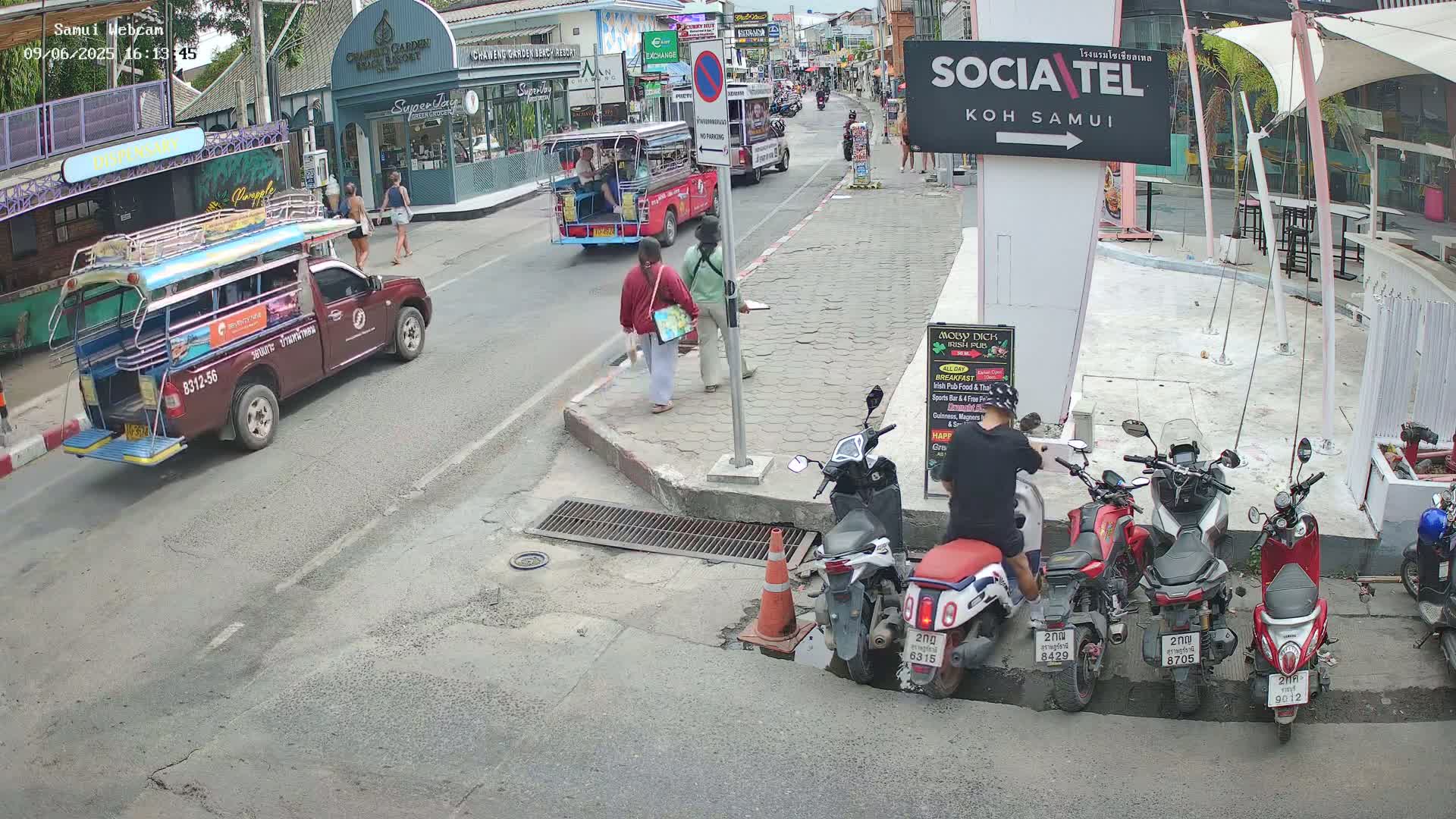 A street scene on a sunny day shows several people walking and standing near parked motorcycles and a red songthaew, with other vehicles in the distance.