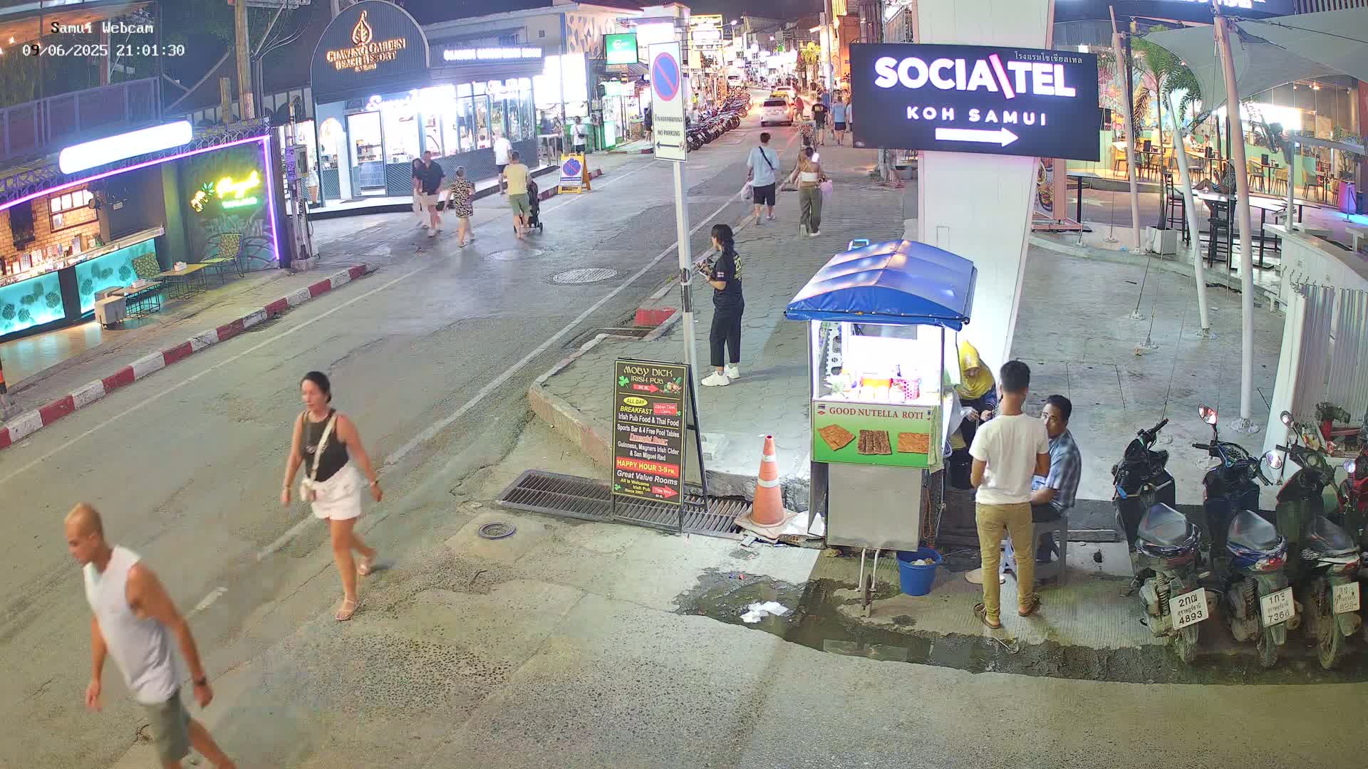 A nighttime street scene shows pedestrians walking past shops and food stalls, with several parked motorbikes visible under outdoor lighting.