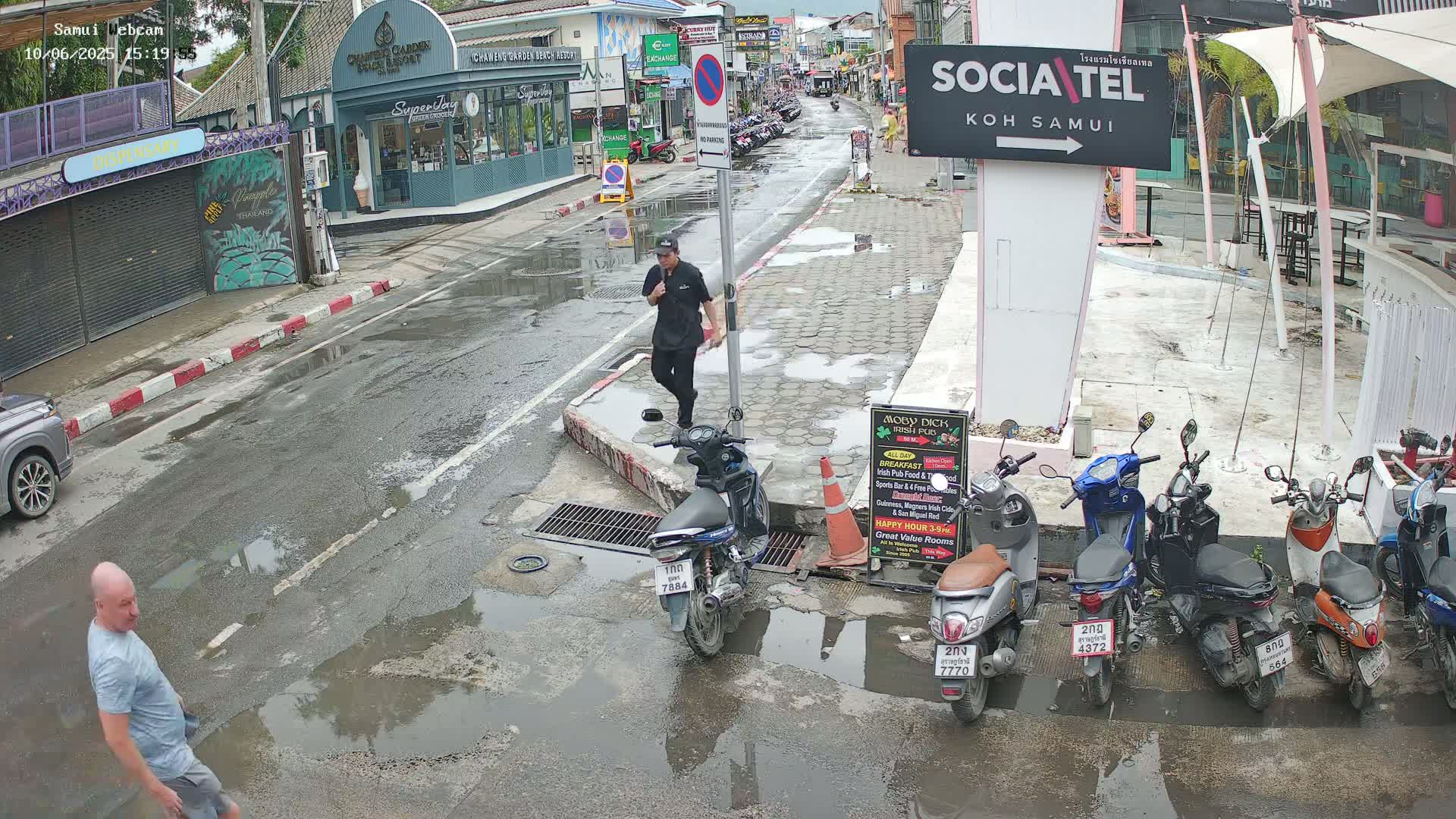 A wet street scene shows several parked motorbikes and a man walking, with a second man further down the street; puddles are visible on the roadway.