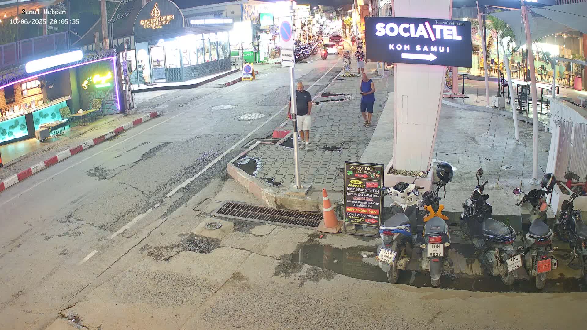 A nighttime street scene shows two pedestrians walking past parked motorbikes and several businesses under dim lighting.