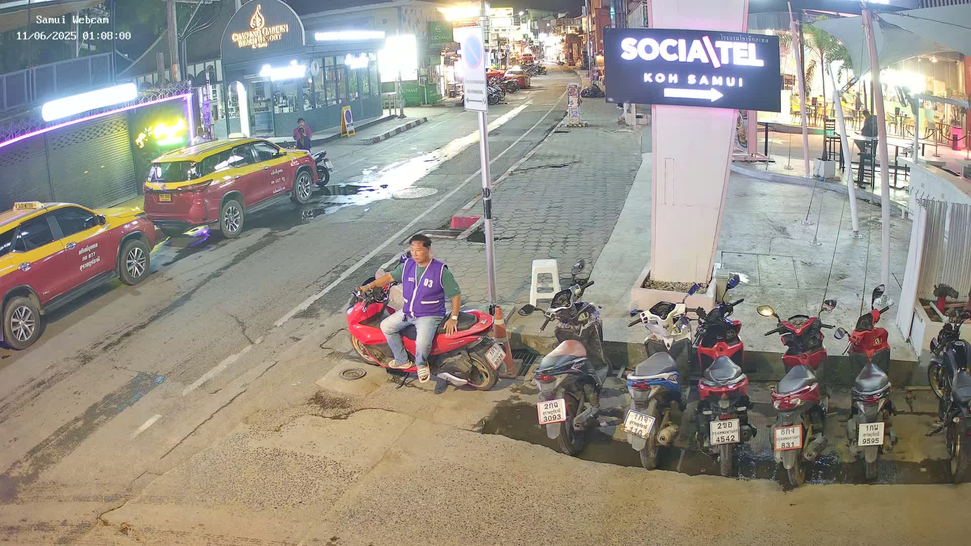 A man sits on a red scooter in a nighttime street scene with parked cars and motorcycles, under a partly cloudy sky.