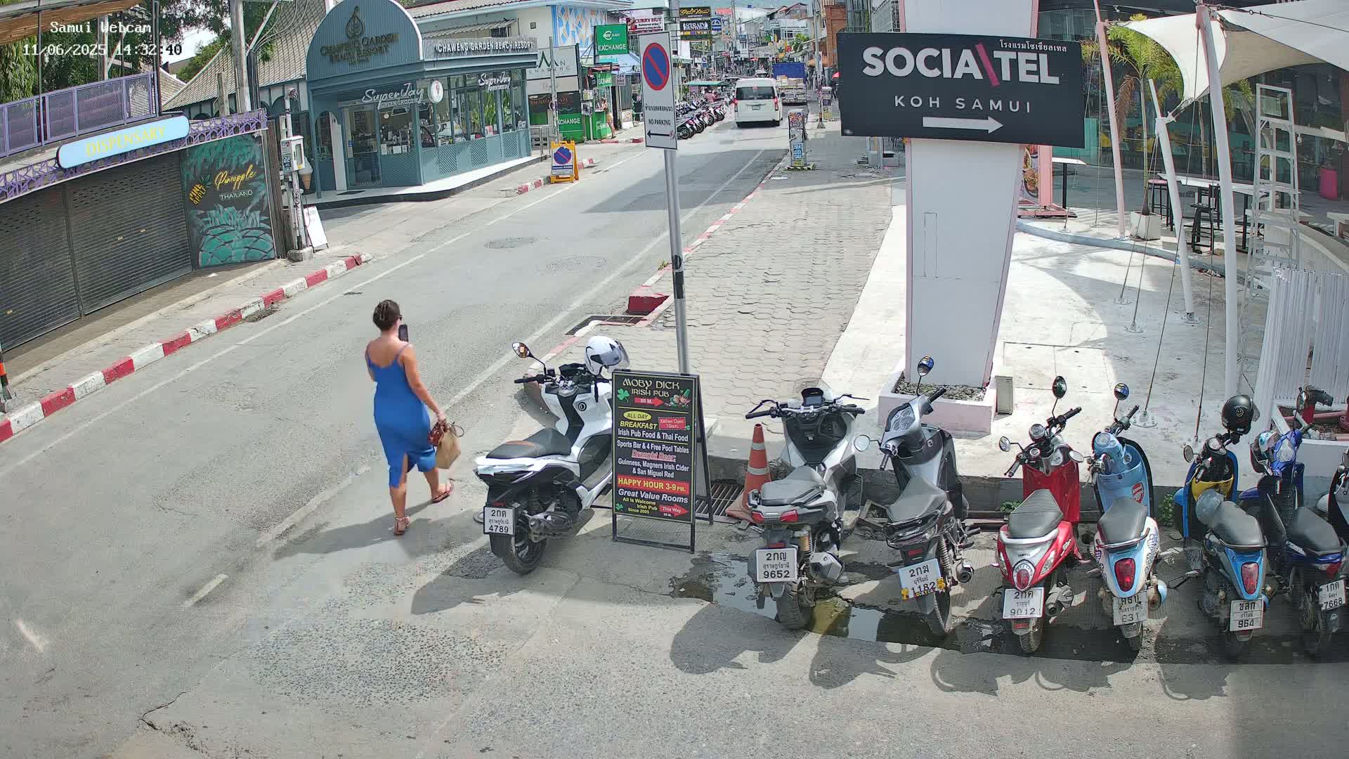 A woman in a blue dress walks down a sunny street lined with parked motor scooters and shops.