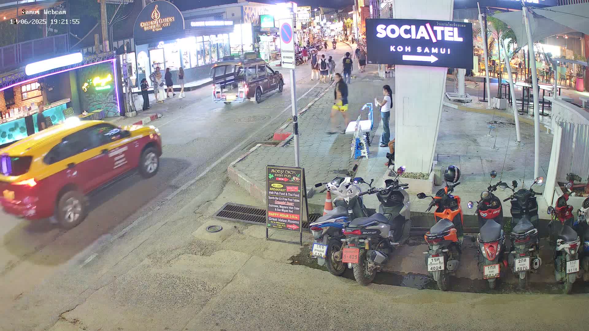 A nighttime street scene shows a yellow and red taxi driving past pedestrians and parked motorbikes under streetlights.