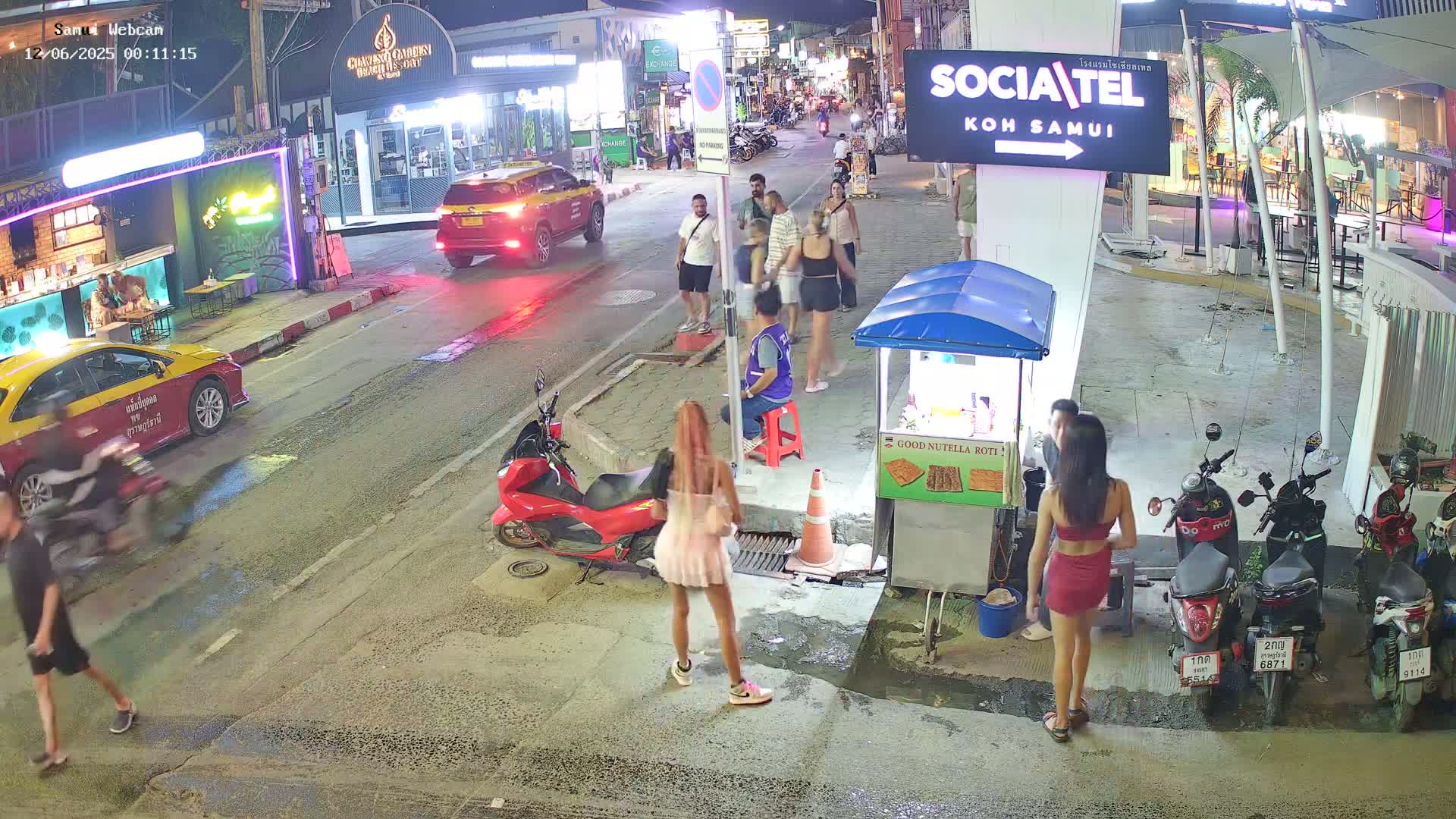 A nighttime street scene shows people walking and standing near parked motorcycles and cars, with a food vendor's cart nearby.