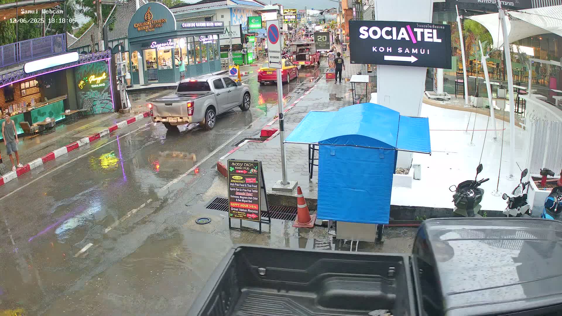A rain-slicked street scene shows several vehicles, including a pickup truck, navigating past shops and restaurants, with motorbikes parked nearby under a partly cloudy sky.