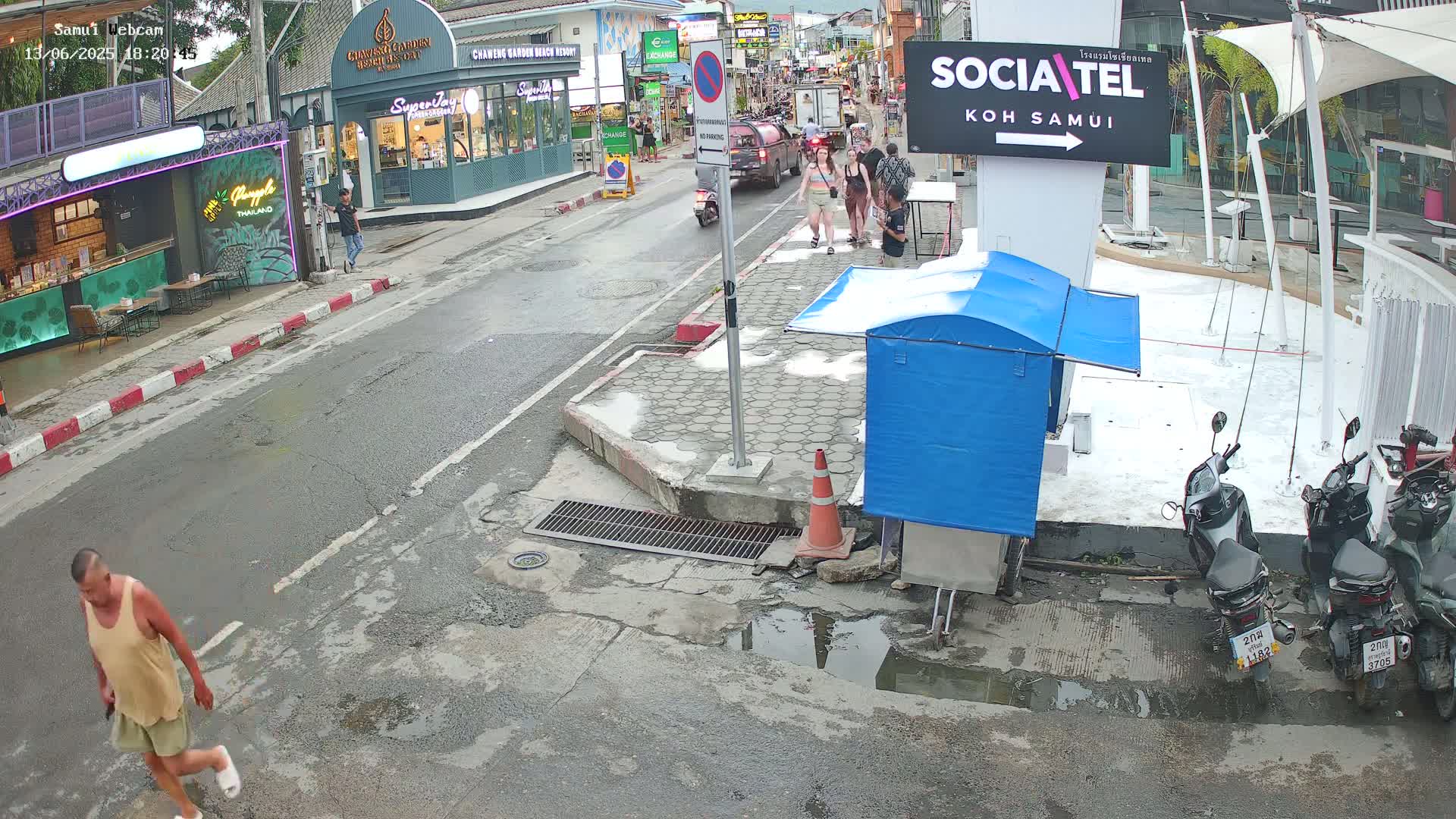 A man walks down a wet street in front of shops and parked motorbikes, with several people standing on the sidewalk.