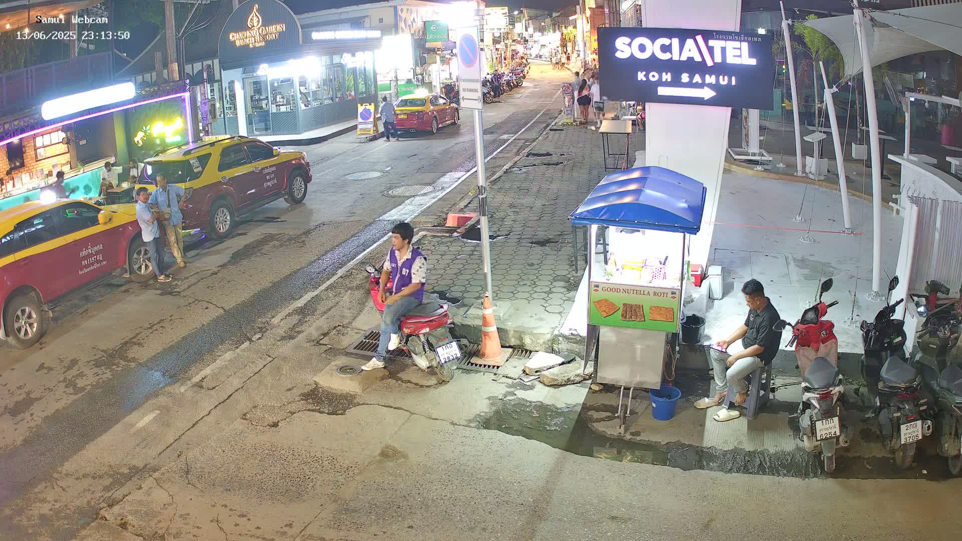 A nighttime street scene shows several people standing and sitting near parked cars and motorcycles, with a food stand and several buildings in the background under clear skies.