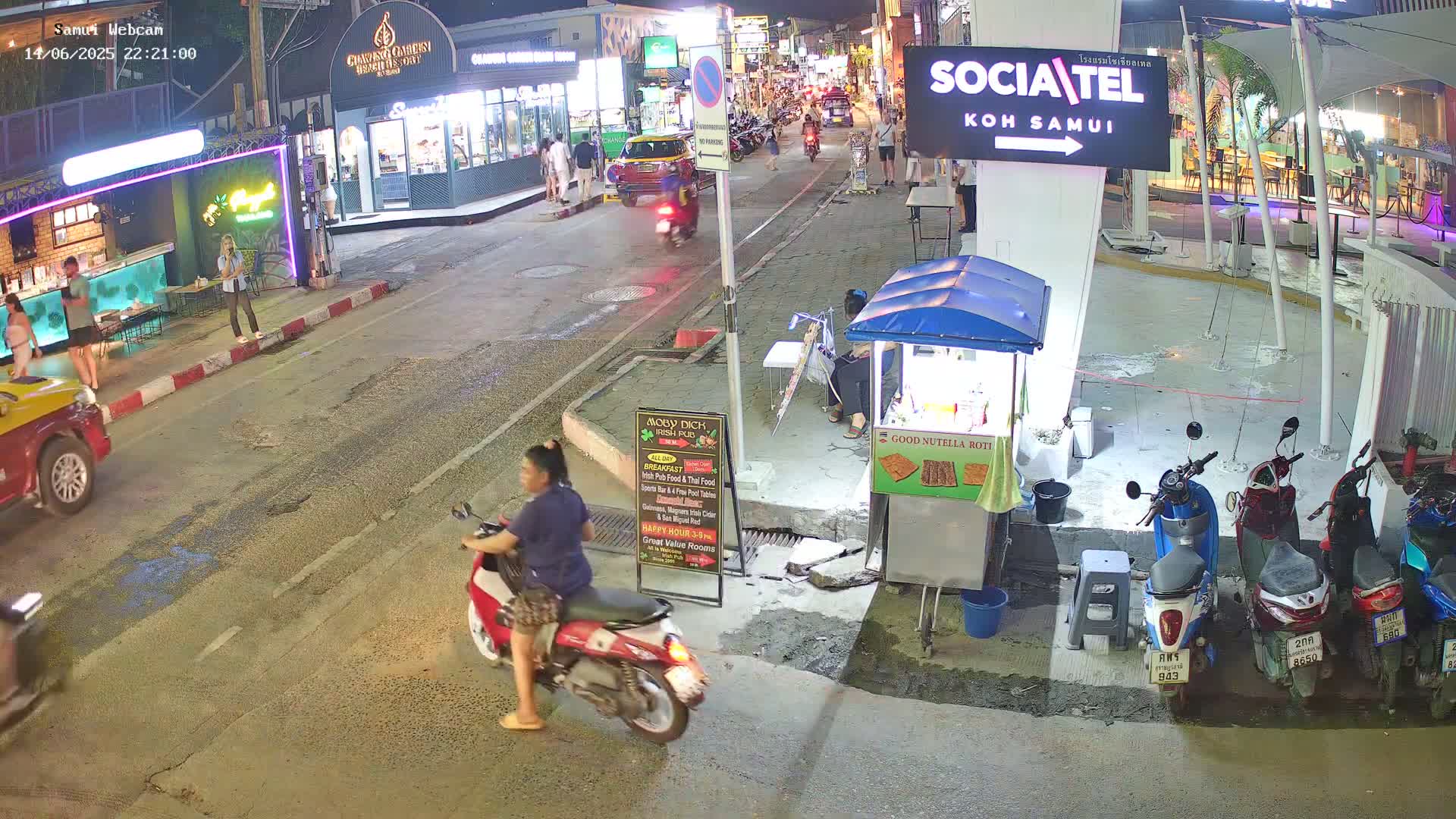 A nighttime street scene shows a person riding a red scooter past a food stand, with several parked motorbikes and pedestrians visible along a road lined with buildings.