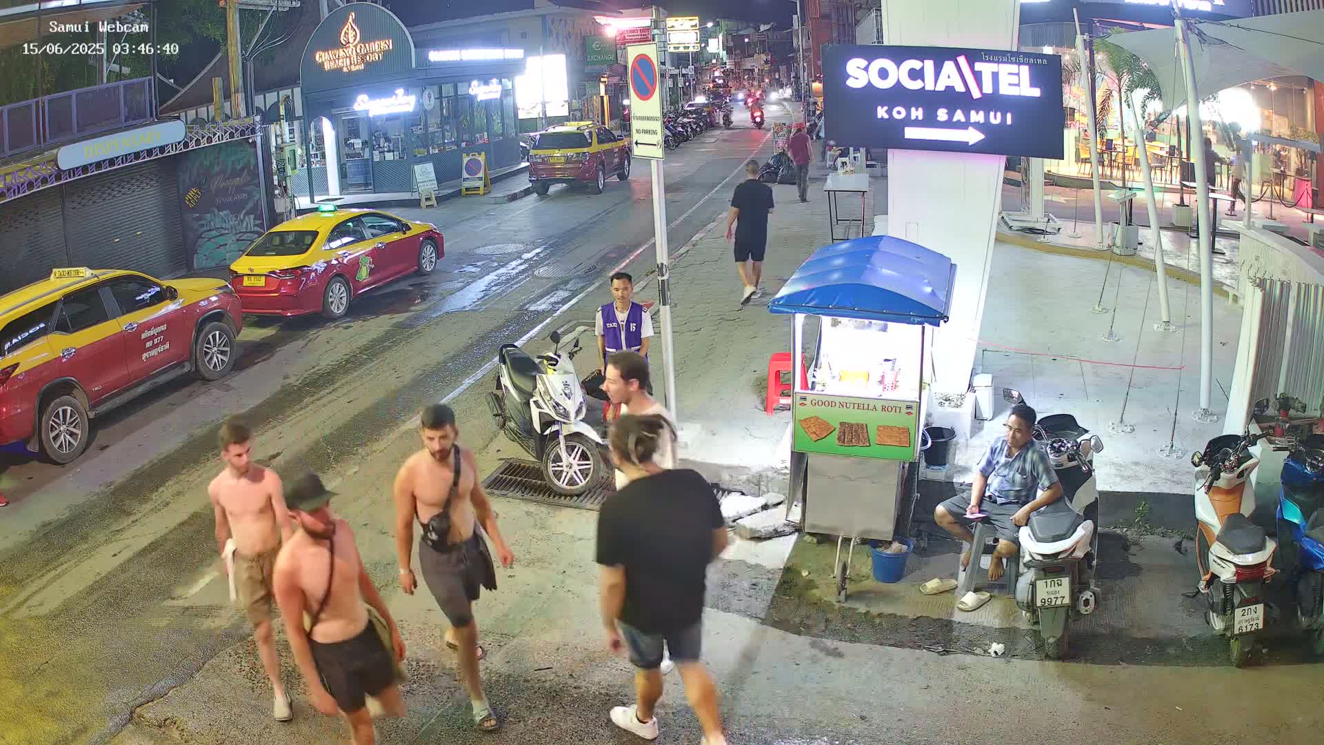 A nighttime street scene shows a person riding a red scooter past a food stand, with several parked motorbikes and pedestrians visible along a road lined with buildings.